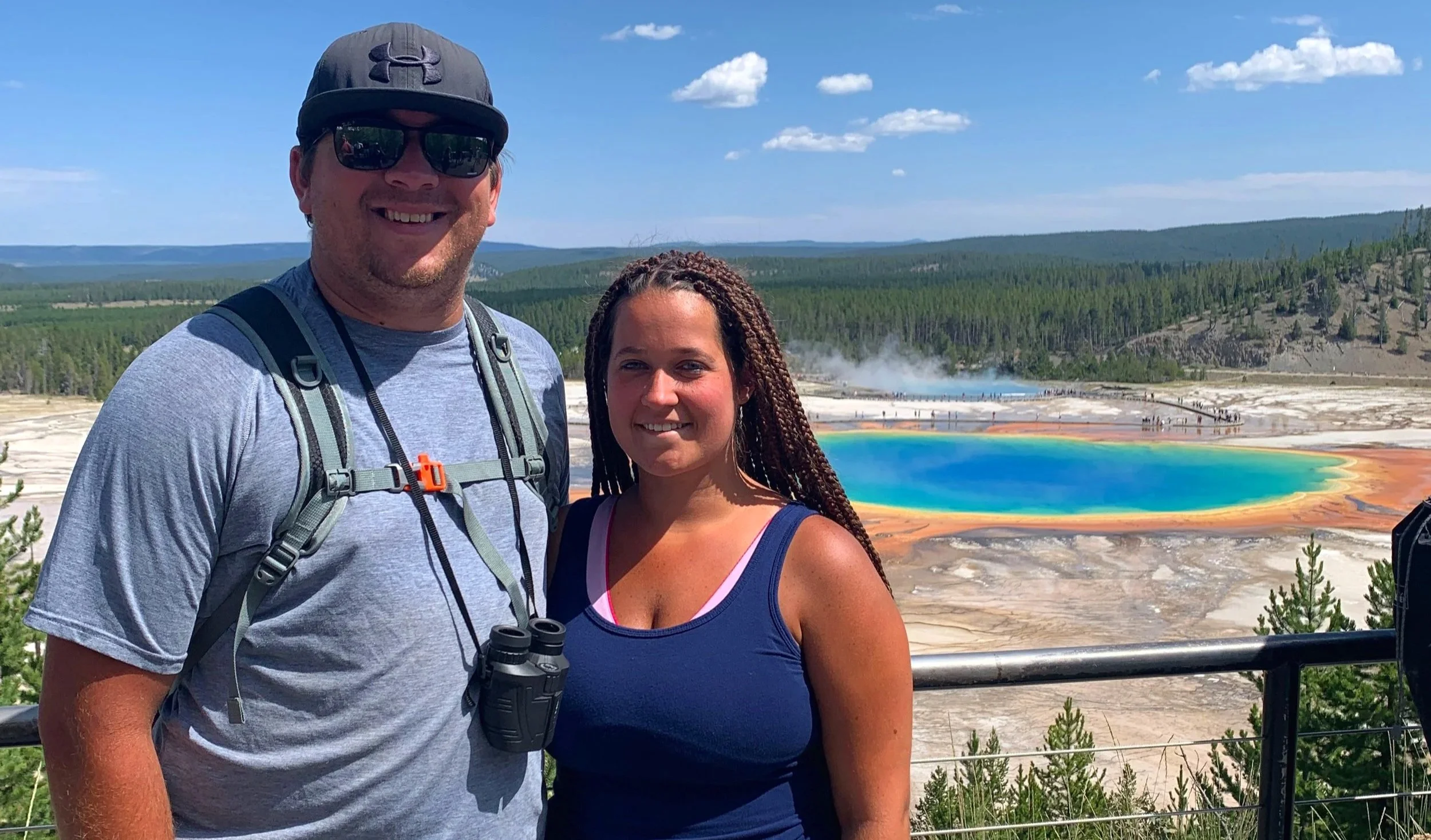 A couple posing in front of the Grand Prismatic Spring from a higher vantage point, showcasing the vibrant colors of the geothermal feature and surrounding landscape.