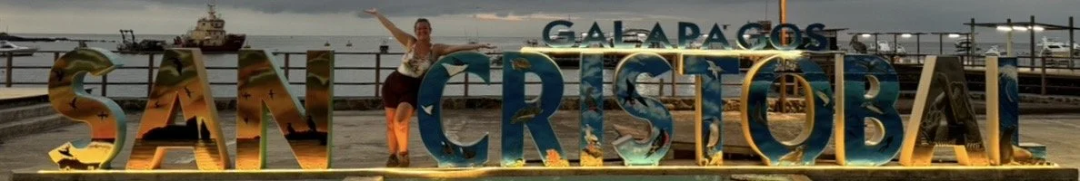 A young woman standing in front of the San Cristóbal sign in the Galápagos Islands at night, illuminated by lights.