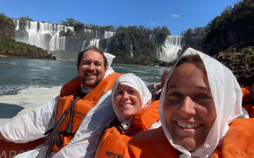 Three people on a boat at Iguazu Falls, Brazil, surrounded by cascading waterfalls and mist.