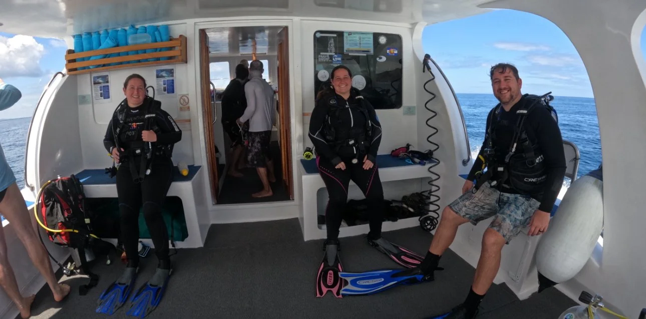 Three people sitting on a boat in the Galápagos Islands, wearing scuba gear and preparing to dive.