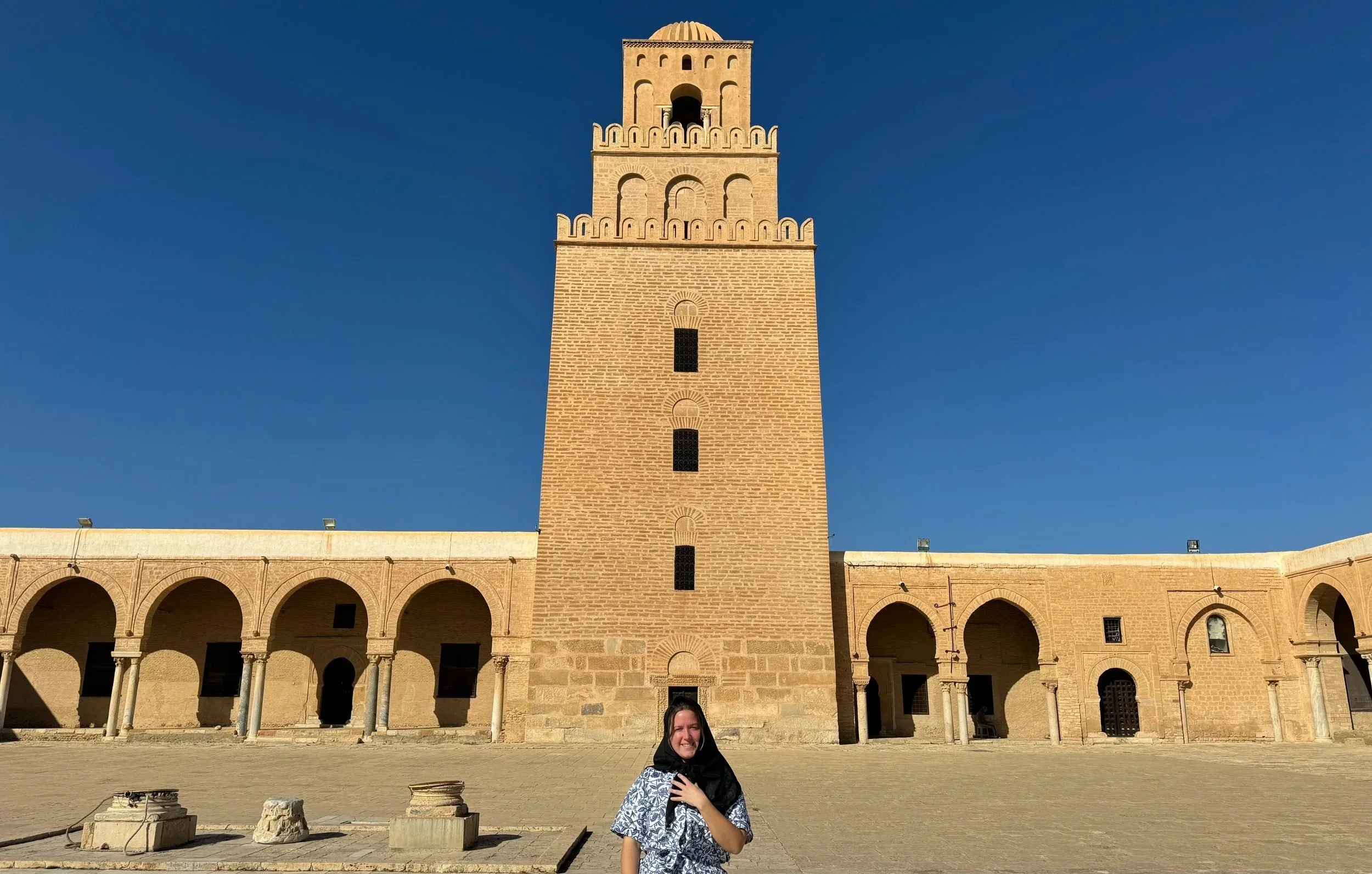 A woman wearing a headscarf standing in front of the Great Mosque of Kairouan in Tunisia, with the mosque’s historic stone architecture visible behind them.