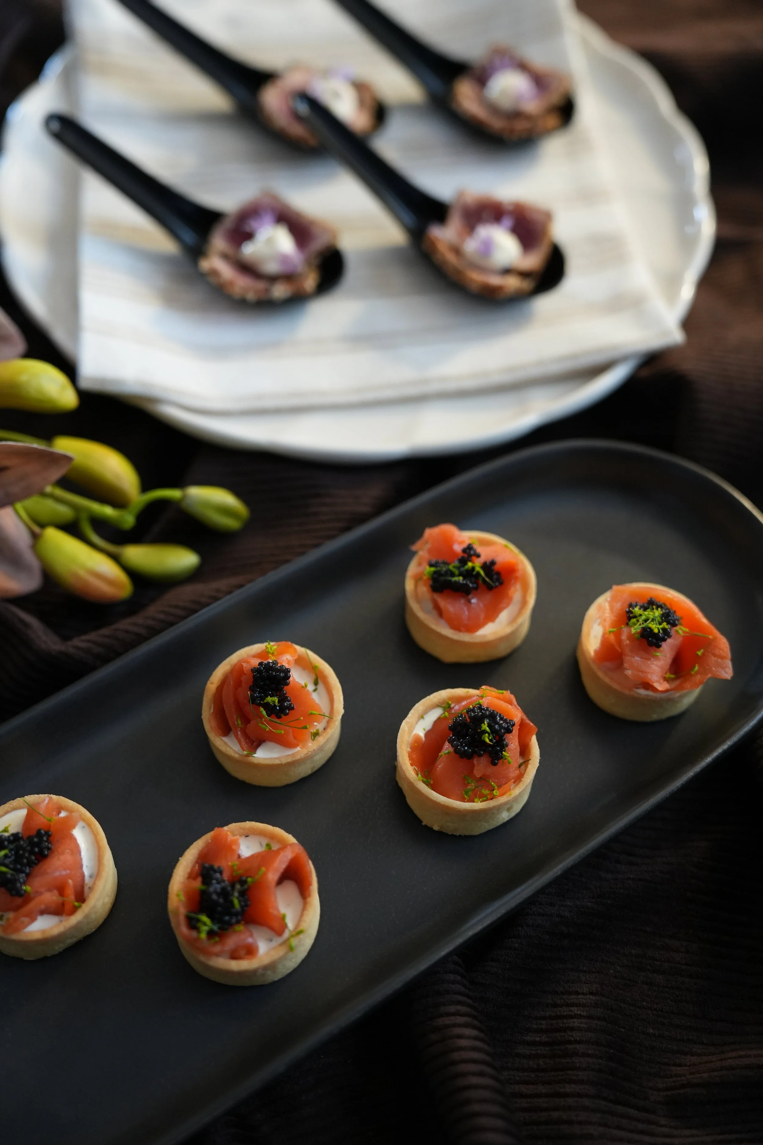 Small canapé appetizers topped with smoked salmon, garnished with black caviar and green herbs, served on a black rectangular plate.