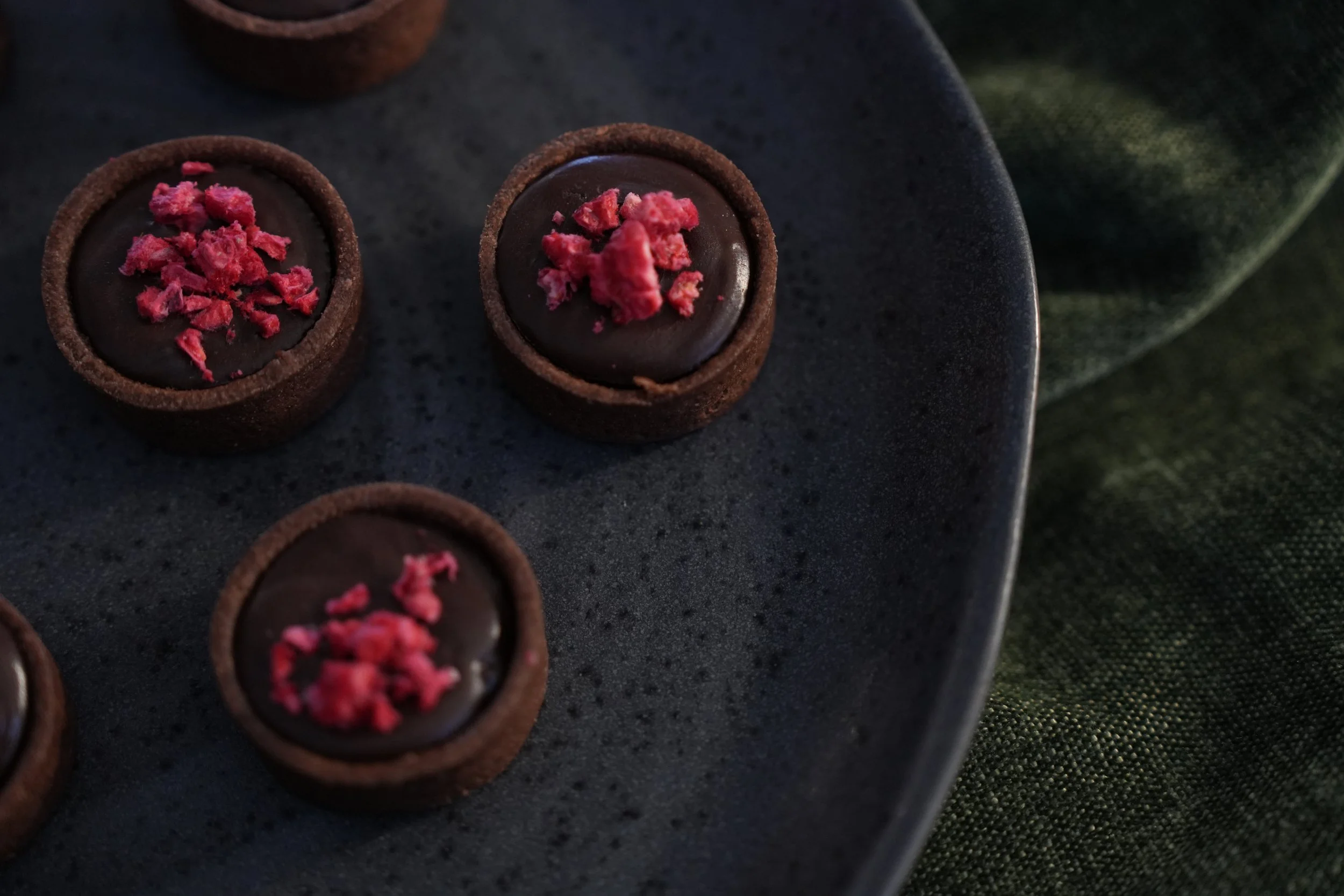 Chocolate tartlets topped with pink crumbles on a dark plate.