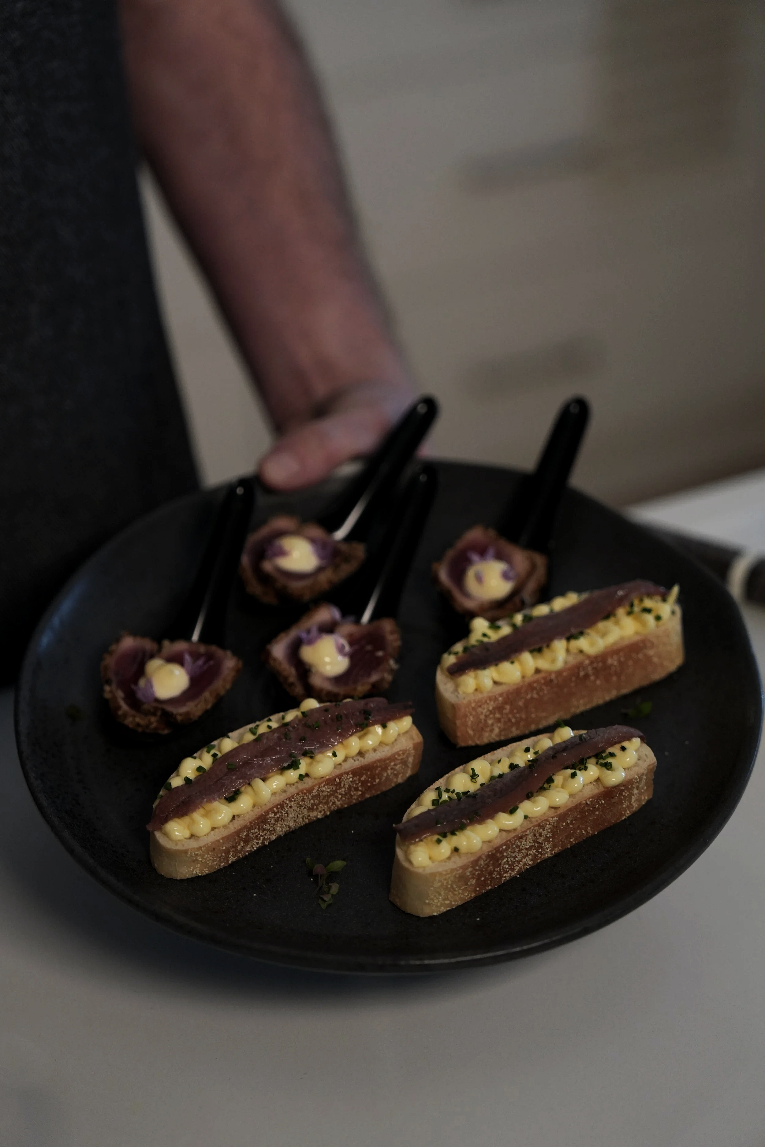 A black plate with three slices of baguette topped with cheese and slices of cured meat, and four small bites of seared meat and sauce held by black forks.