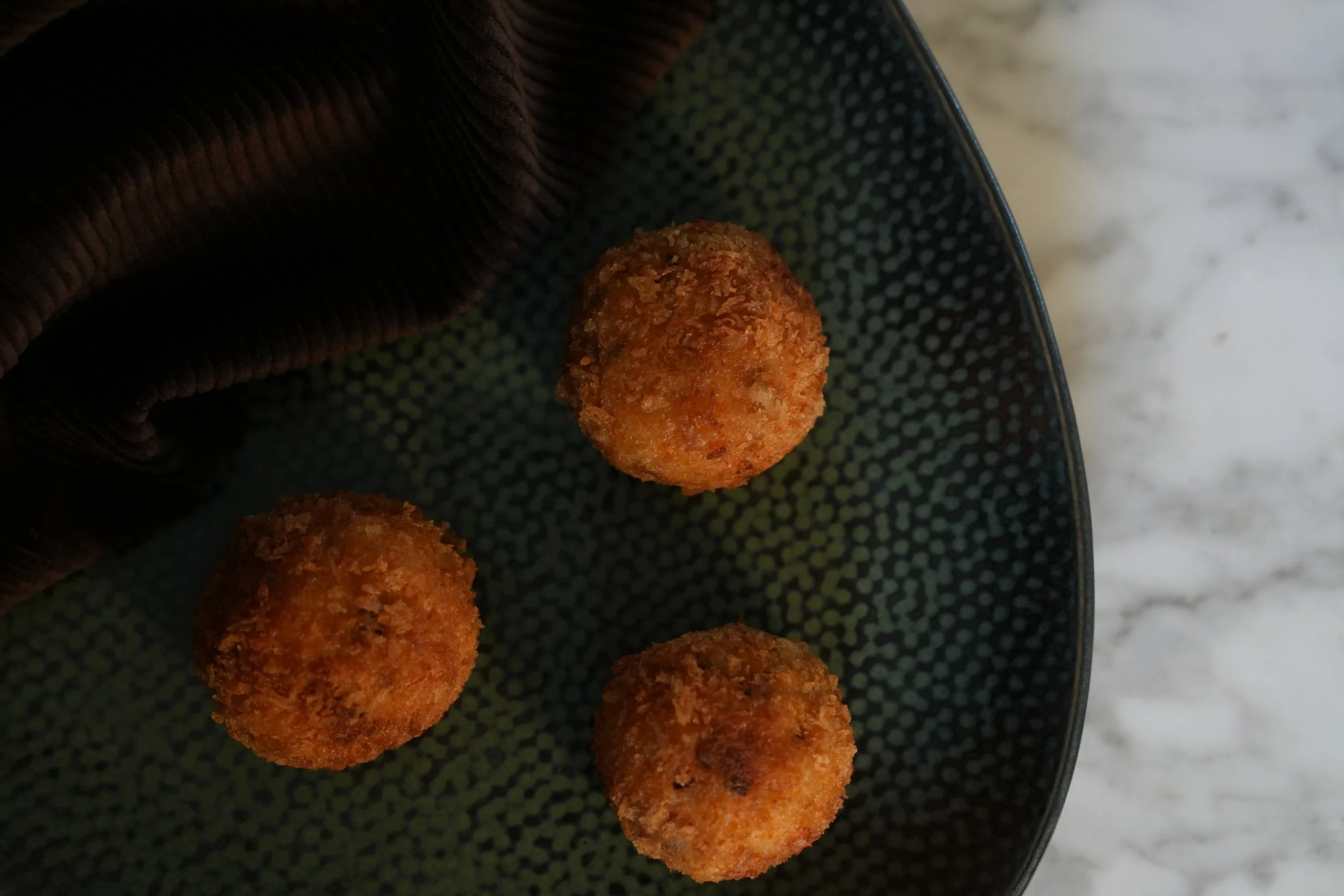 Three fried arancini on a black textured plate with a dark cloth partially covering the plate, placed on a light marble surface. Premium catering for events and corporate functions Sydney