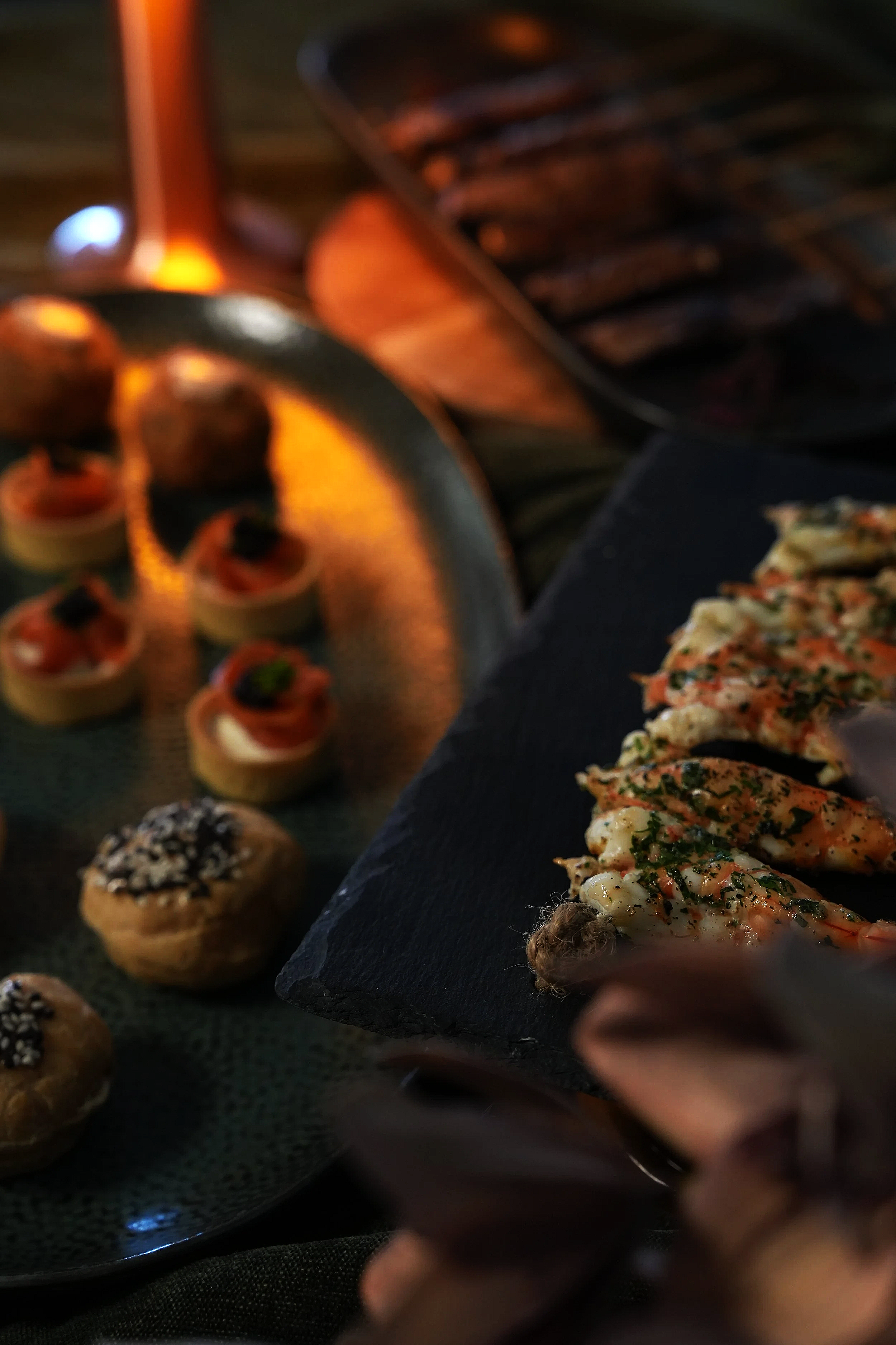 Close-up view of various appetizers at a dimly lit dining setting, including small pastries with toppings and a sliced seafood dish garnished with herbs on a black slate serving platter.