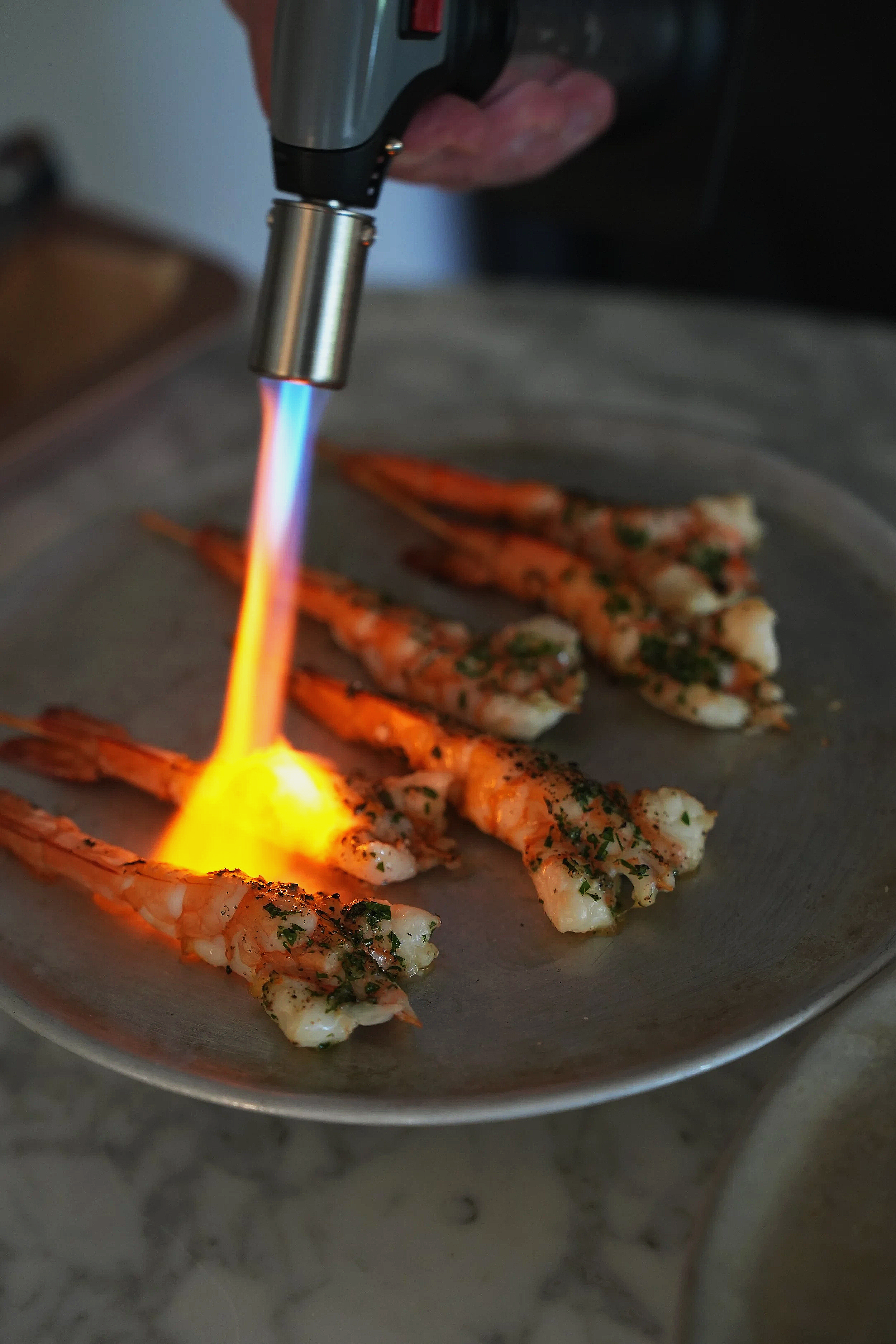 Shrimp being cooked with a culinary torch on a metal plate, garnished with herbs.