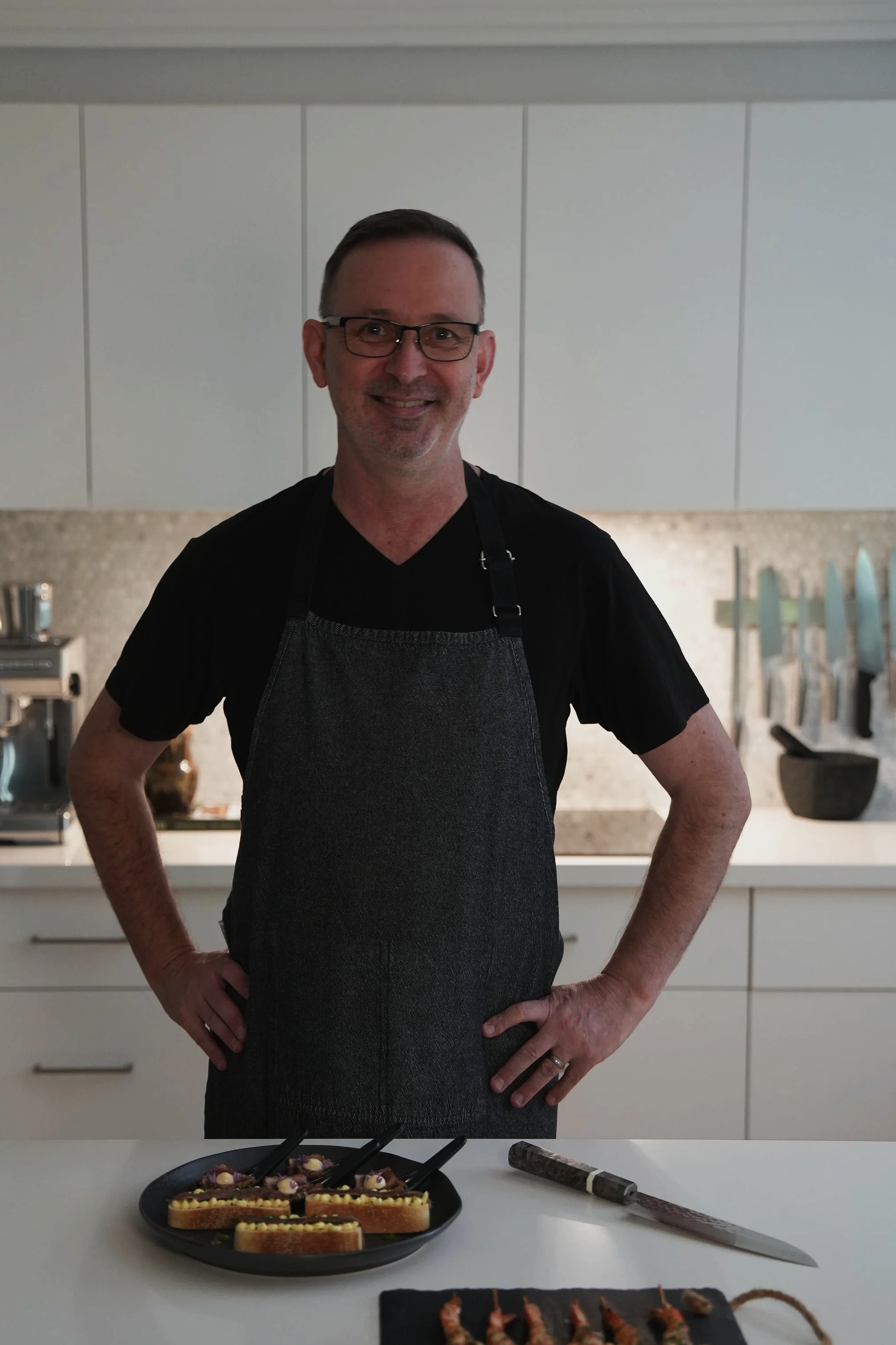 A smiling man wearing glasses, a black shirt, and a dark apron stands behind a kitchen counter with prepared appetizers and knives. The kitchen has white cabinets and a backsplash with a speckled pattern.