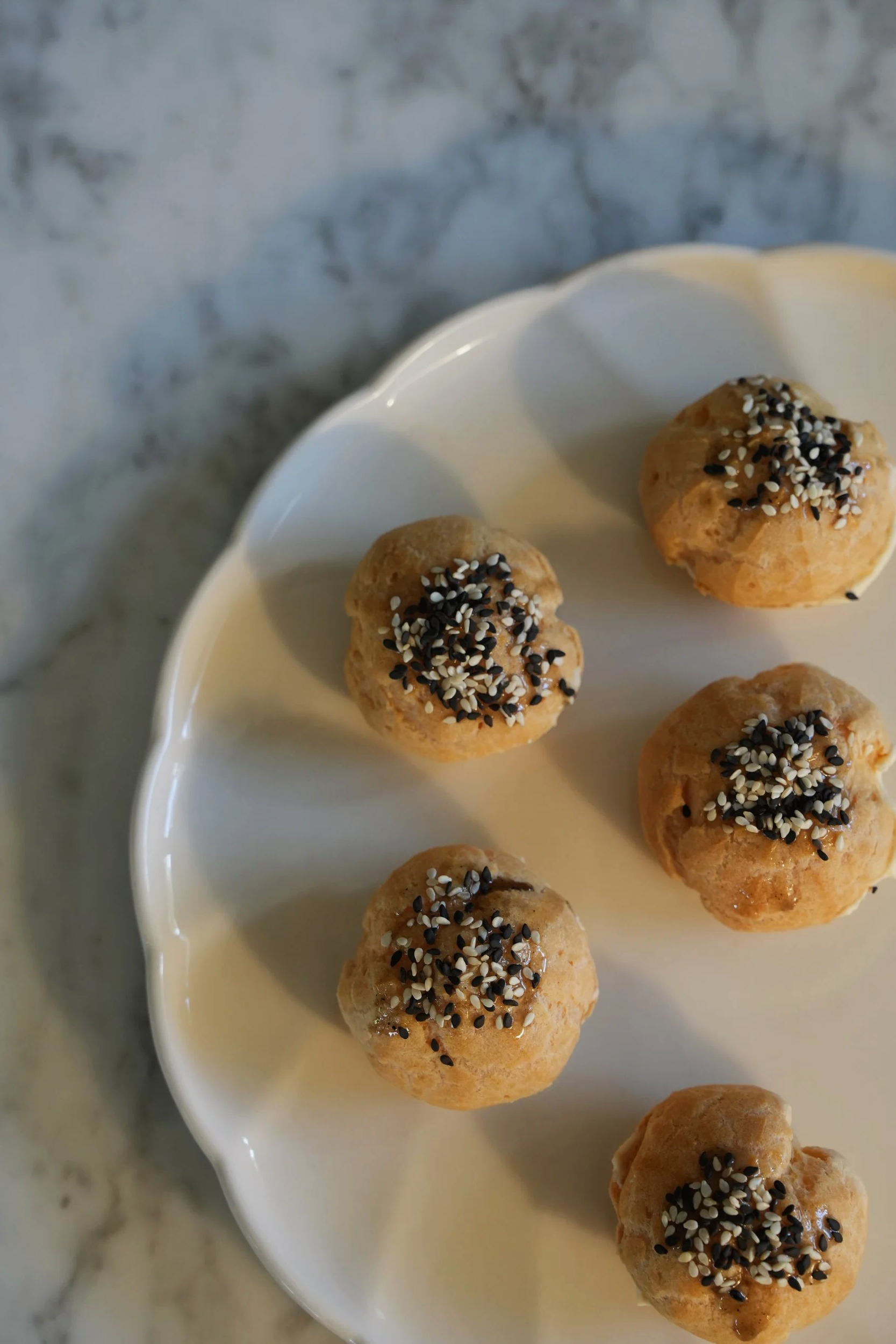 Six round pastry balls topped with black and white sesame seeds on a white scalloped plate, against a marble surface background.