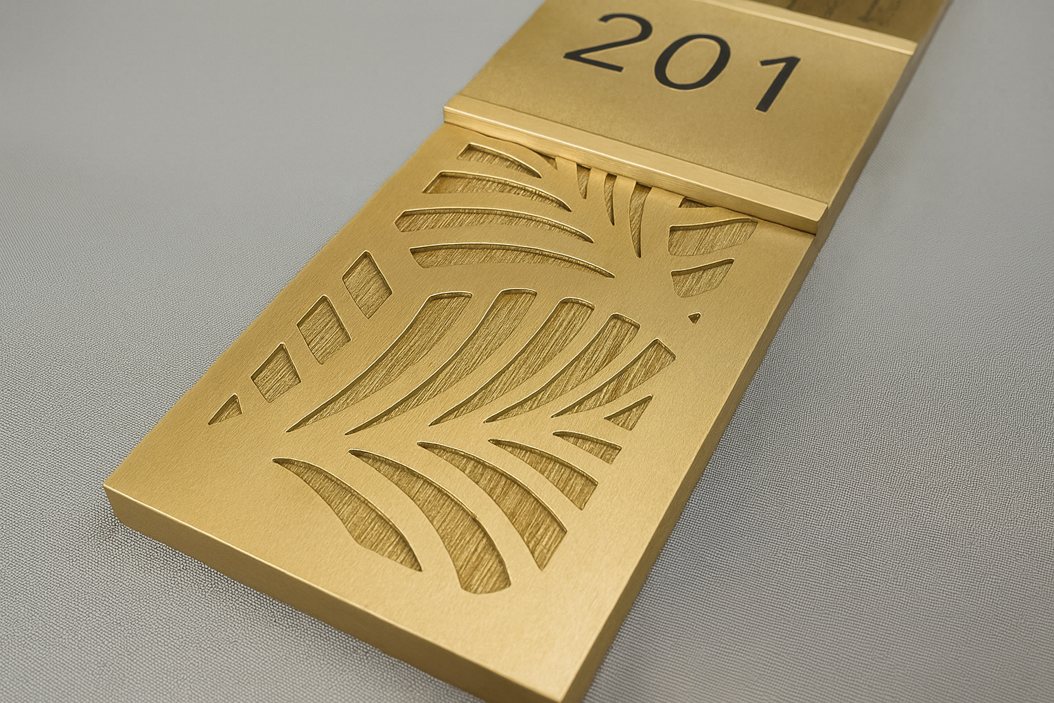 Close-up of a modern custom brass sign panel with intricate cut-out leaf patterns. The brushed brass surface has a warm golden tone and a matte finish, showcasing precision craftsmanship and elegant design on a neutral gray background.