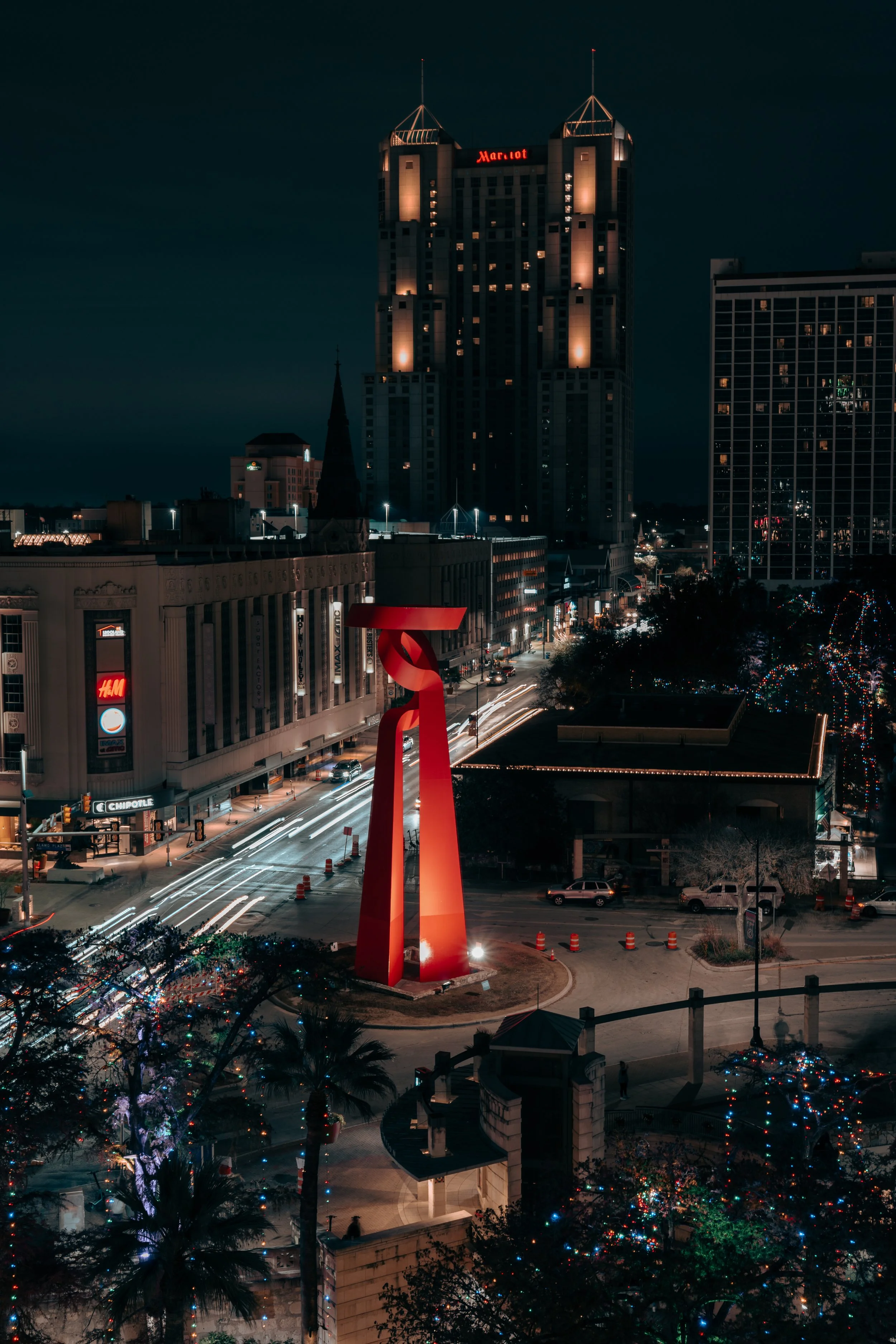 Nighttime cityscape of Downtown San Antonio, Texas. Bustling city streets with friendly tourists, moving vehicles, decorated with Christmas lights, and a large red sculpture in the foreground.