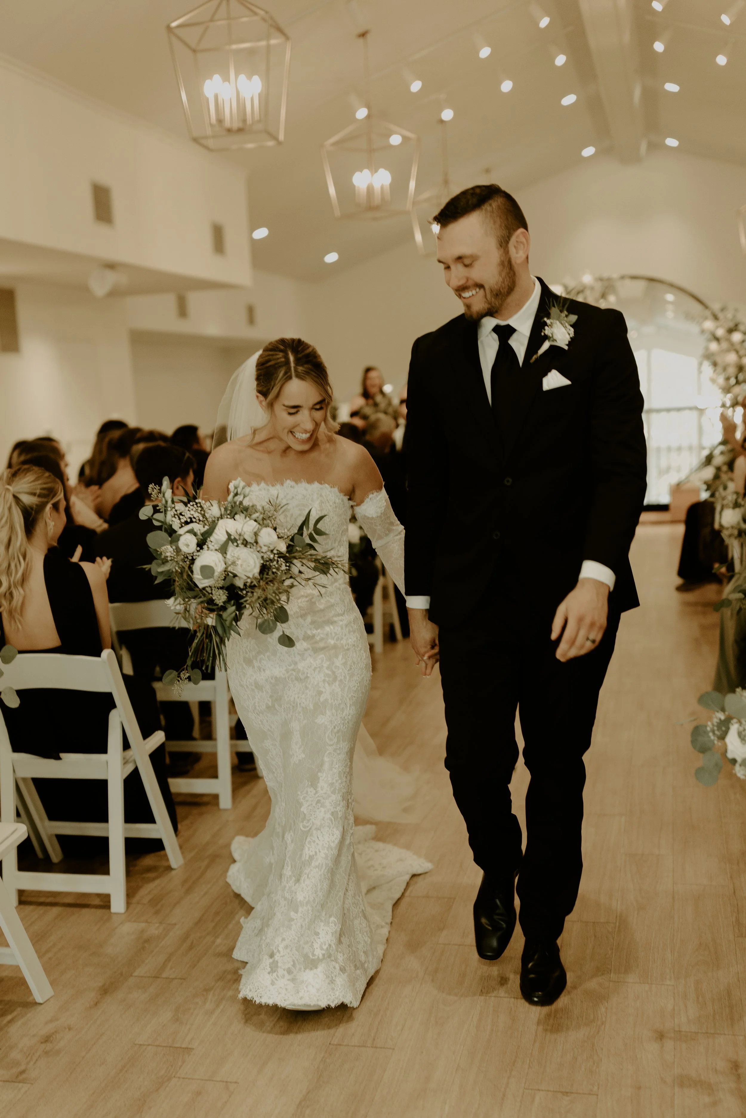 Bride and groom walking down the aisle at their wedding ceremony, surrounded by seated guests, in a decorated indoor venue.
