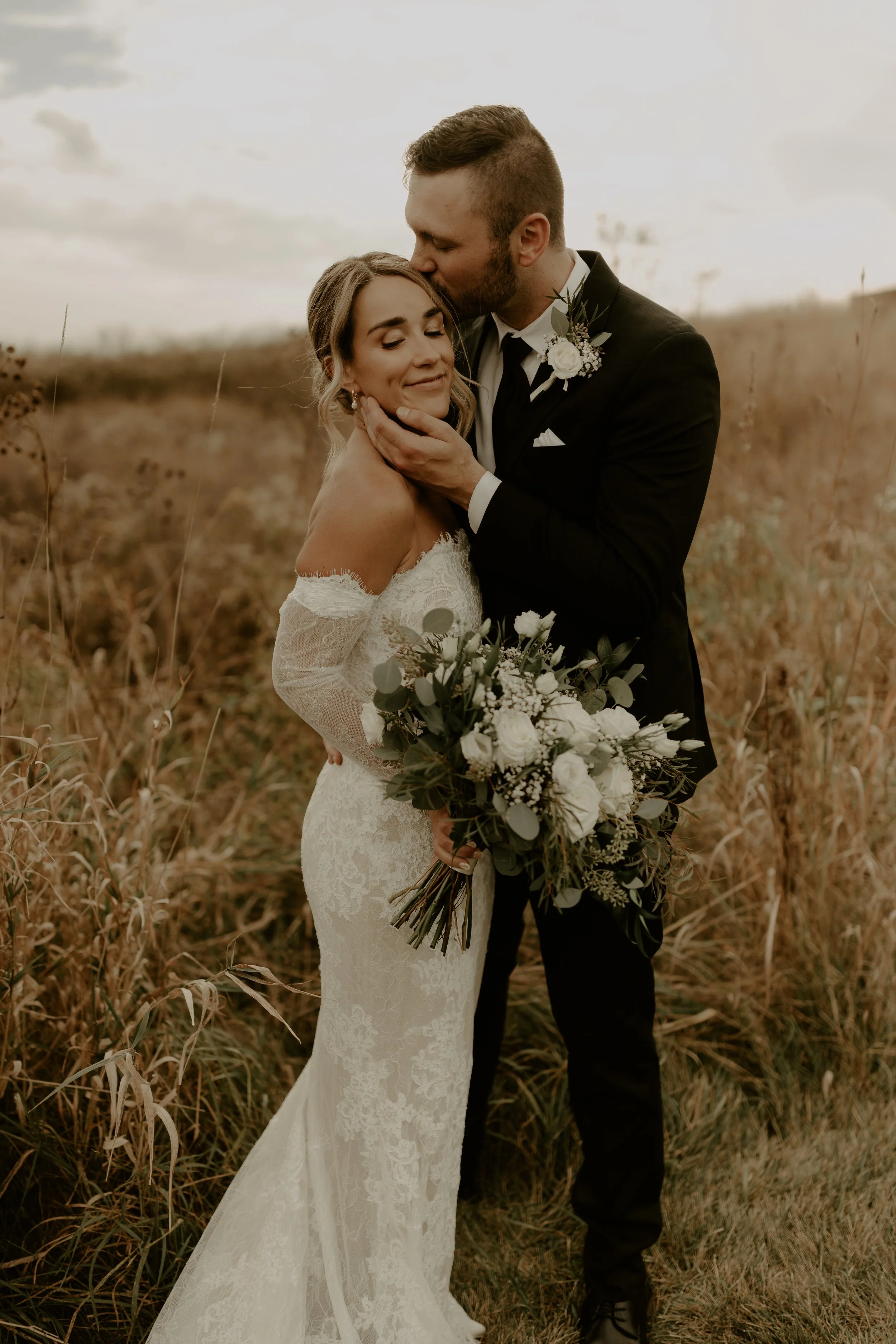 A bride and groom embracing outdoors. The groom is kissing the bride's forehead. The bride holds a bouquet of white flowers and greenery. The bride wears a lace off-shoulder wedding dress, and the groom is in a black tuxedo with a boutonnière. They a