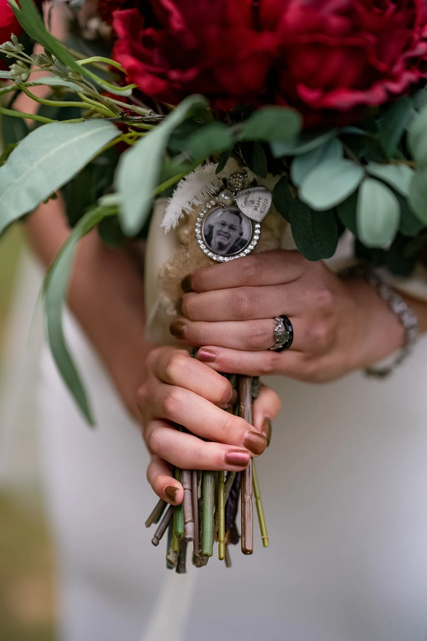 A bride holding a bouquet with a black and white photo charm, greenery, and red flowers, wearing a wedding ring and bracelet.