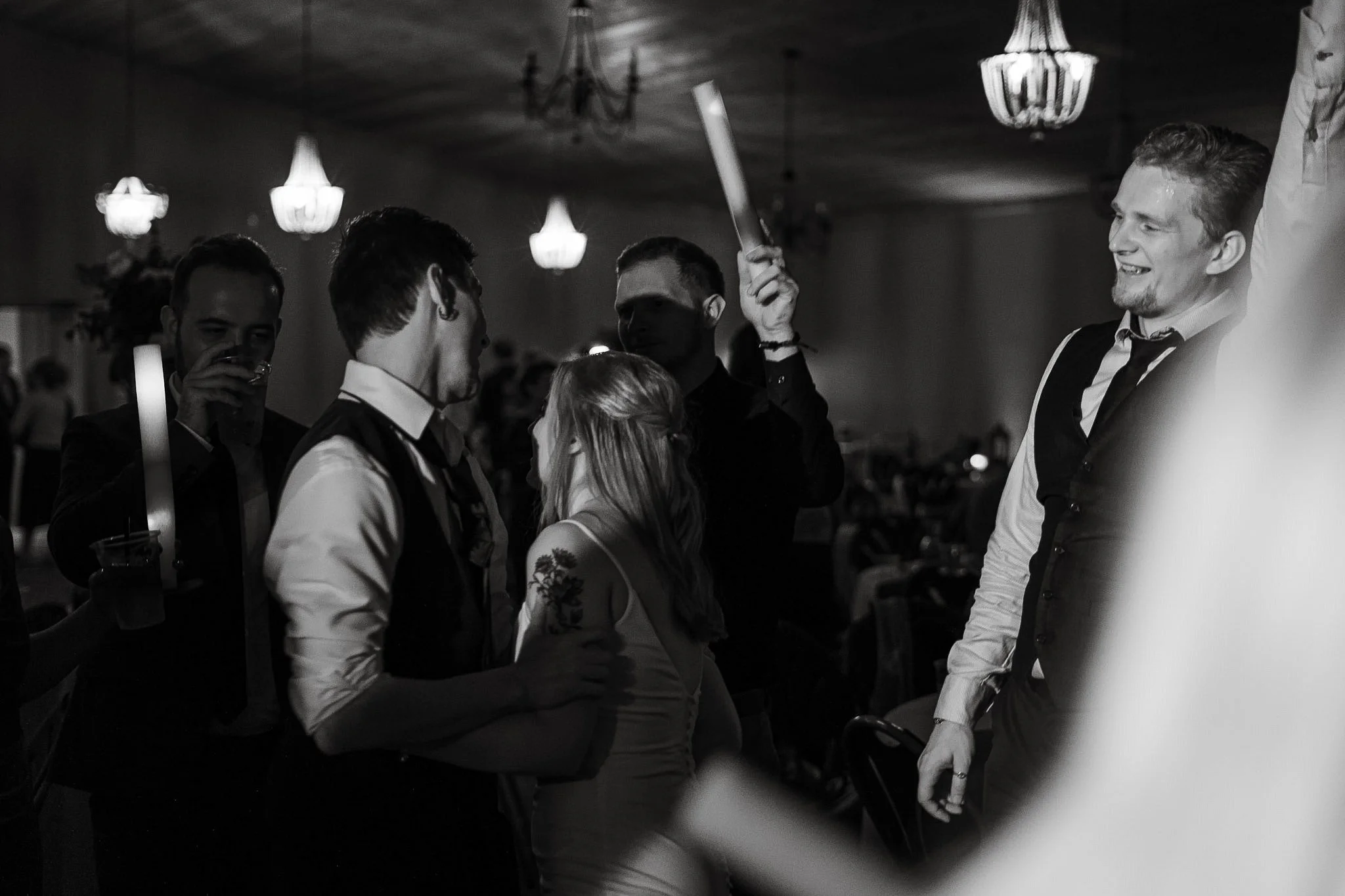 Group of people dancing and laughing at a party or celebration in a dimly lit room with chandeliers.
