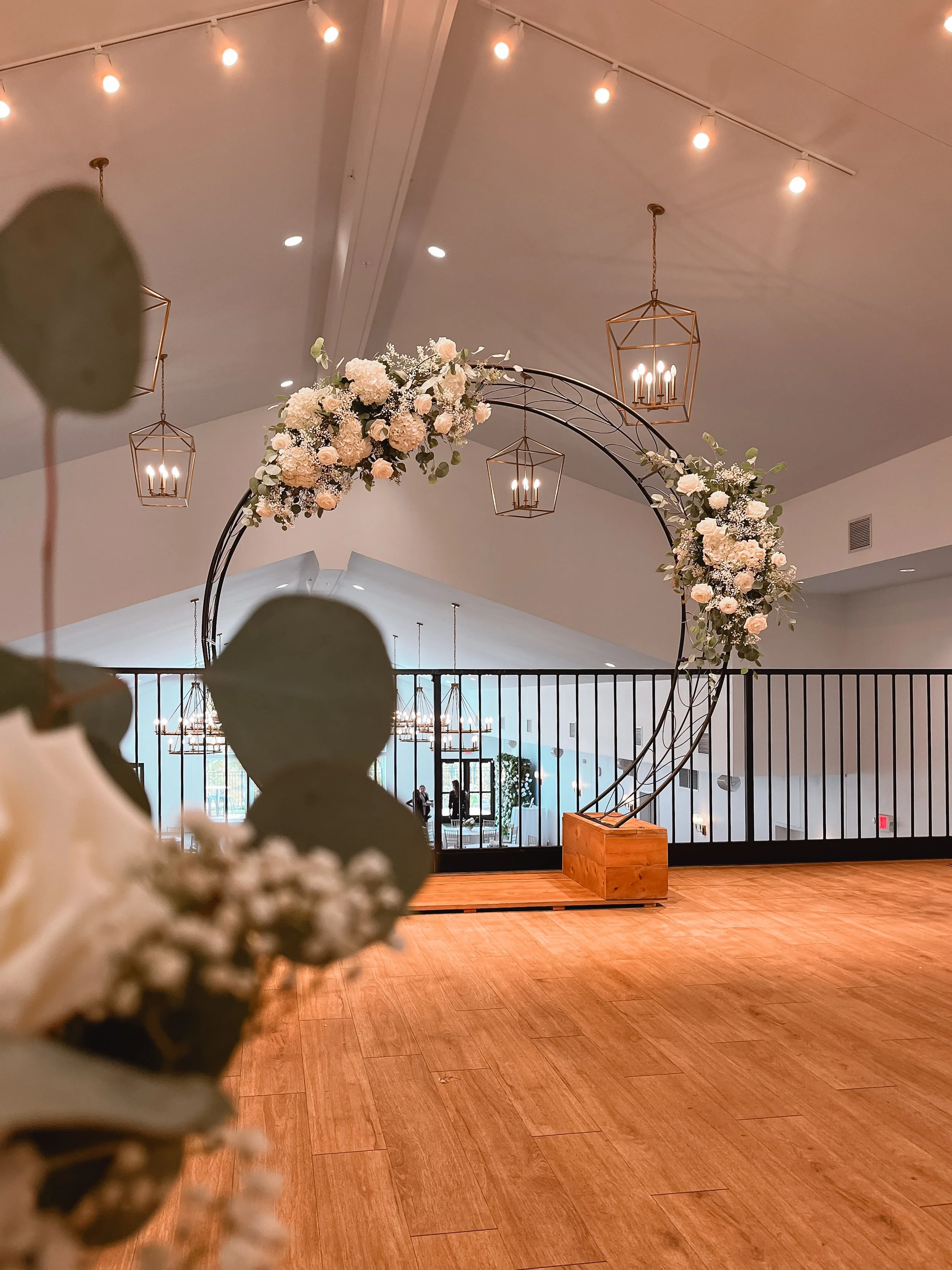 Elegant wedding or event venue with a black metal arch decorated with white and blush flowers, illuminated hanging lanterns, and a wooden stand on a wooden floor.