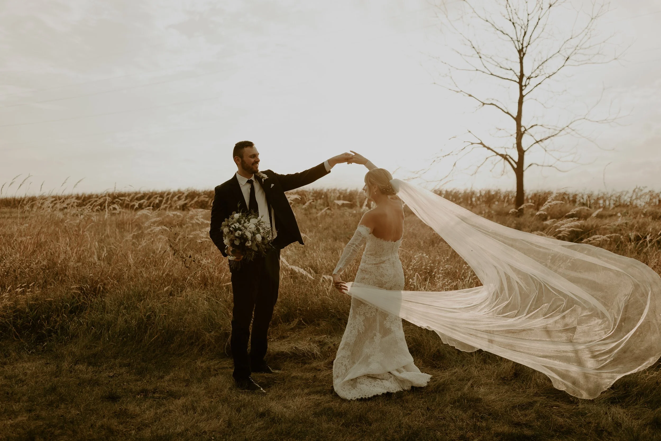 A bride and groom standing in a field, with the groom holding a bouquet and the bride's long veil flowing in the wind, during sunset.