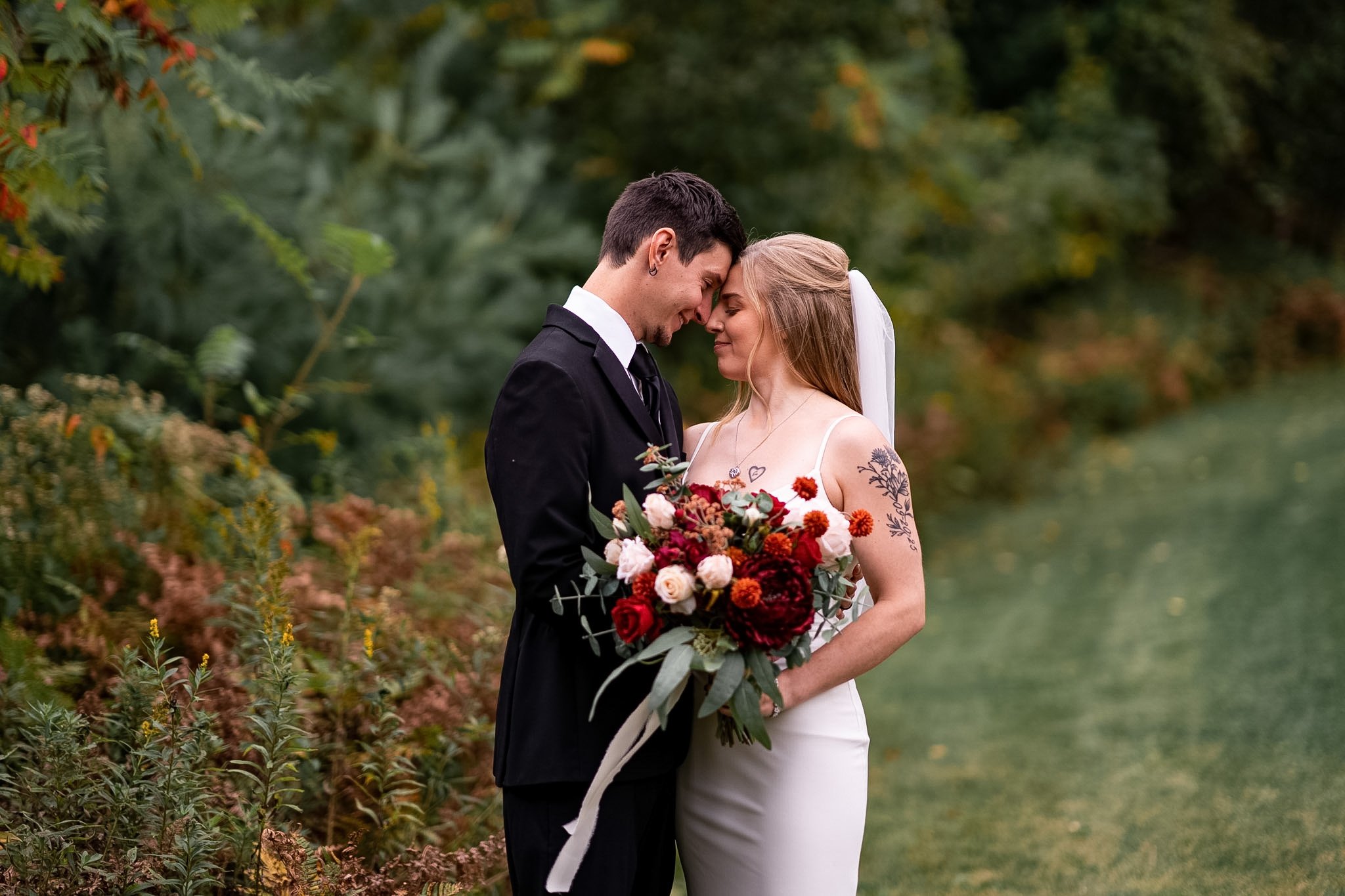 A bride and groom share a tender moment outdoors, with the bride holding a bouquet of red, pink, and white flowers, greenery, and a white ribbon, surrounded by lush greenery.