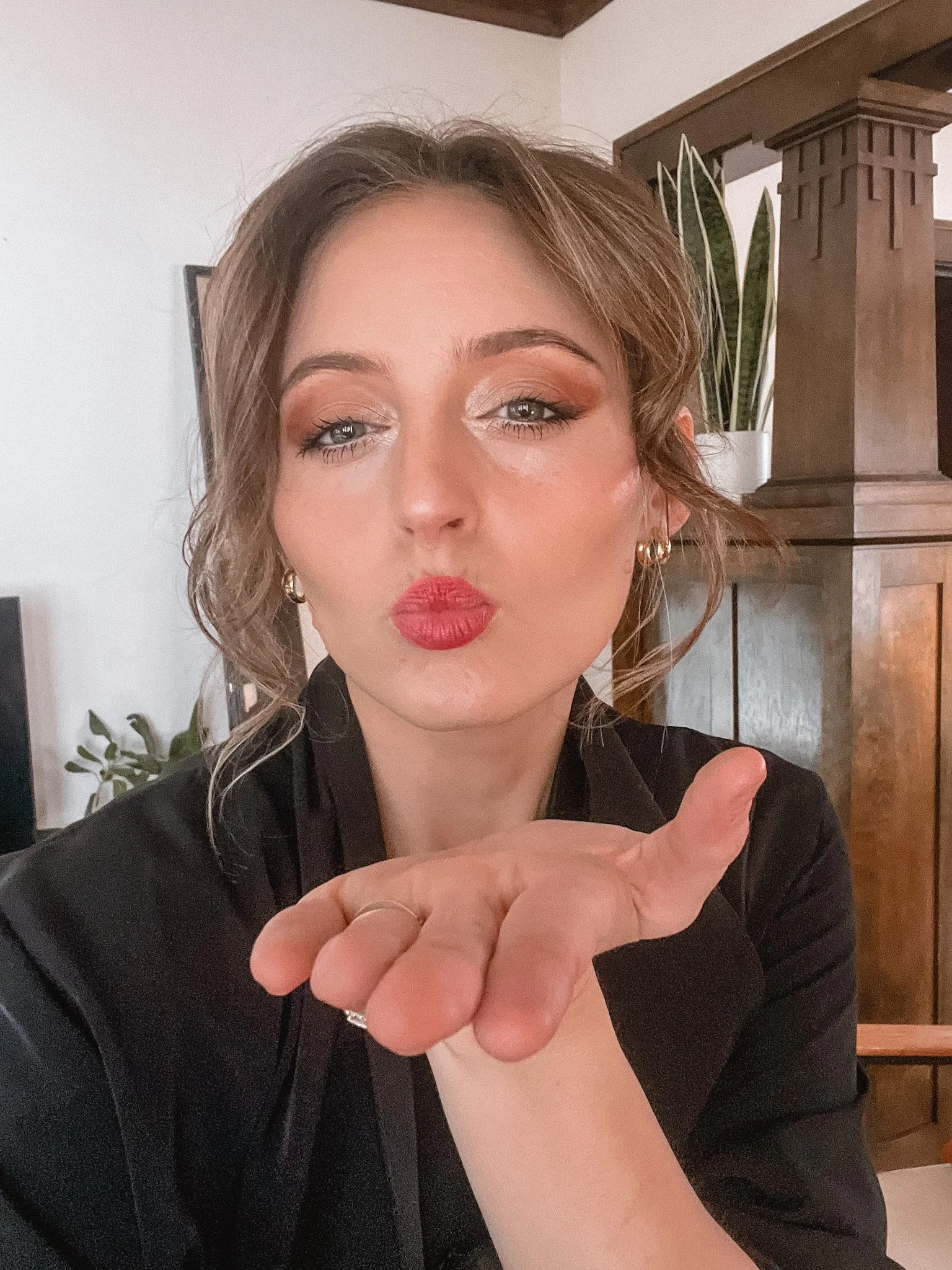 A woman with short, wavy hair and makeup, blowing a kiss while looking at the camera in a room with wooden furniture and potted plants.