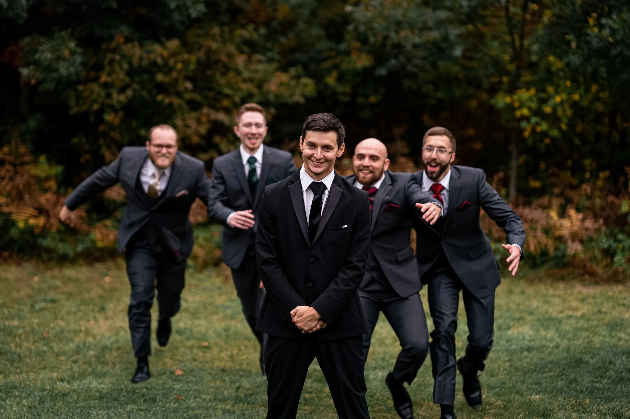 Five men in suits outdoors, one in the front with a smile, others behind making playful gestures.