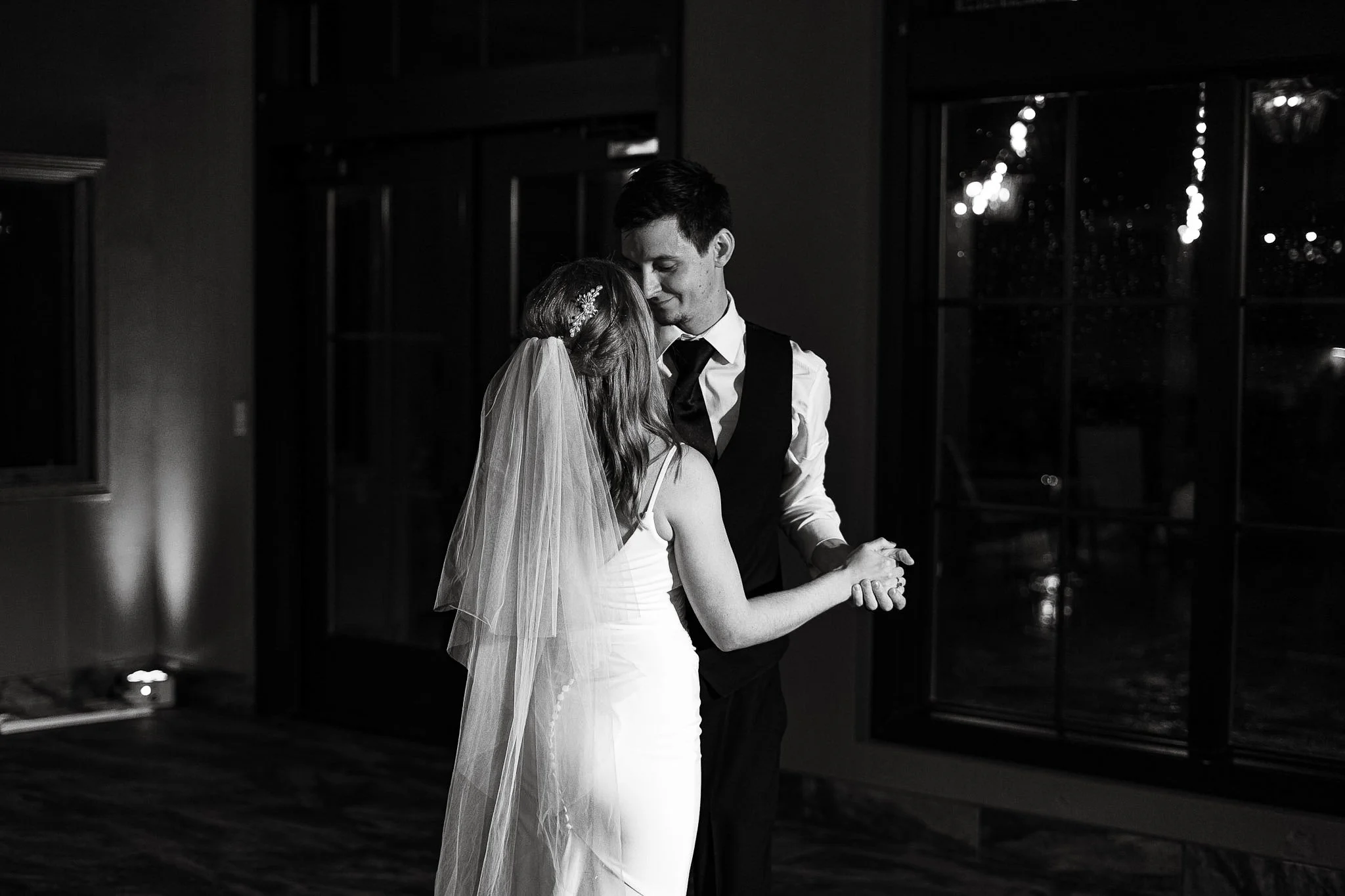 Black and white photo of a bride and groom sharing a dance indoors at night, with a large window showing illuminated outside lights.