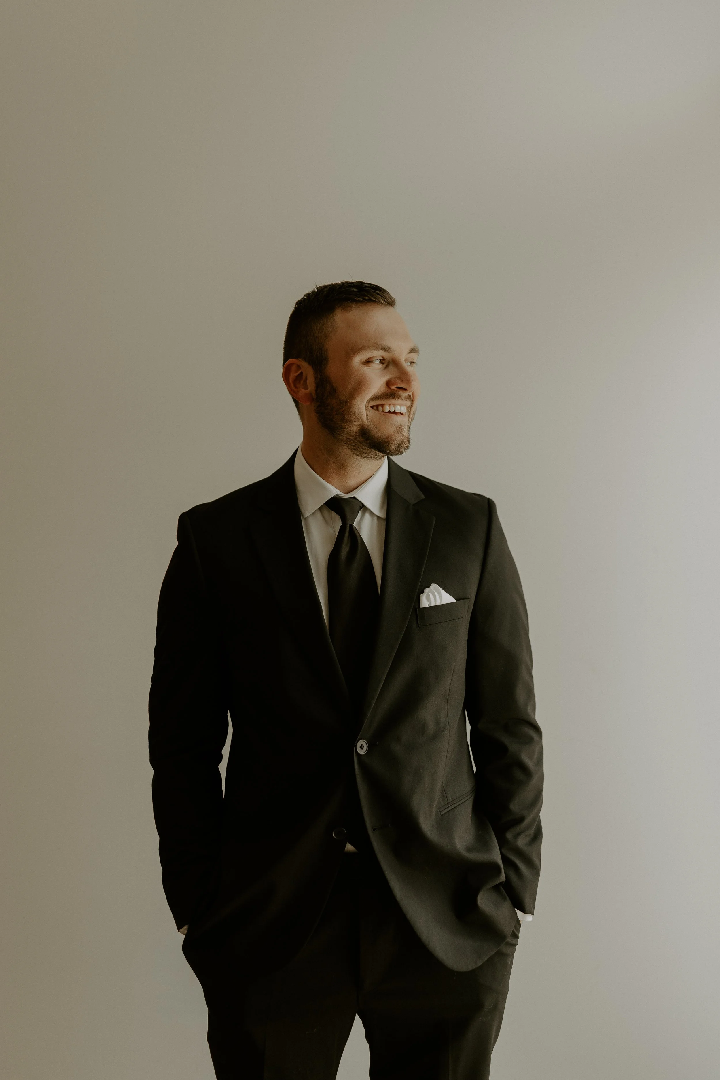 A man in a black suit, white shirt, and black tie standing against a plain light-colored wall, smiling and looking to the side with his hands in his pockets.