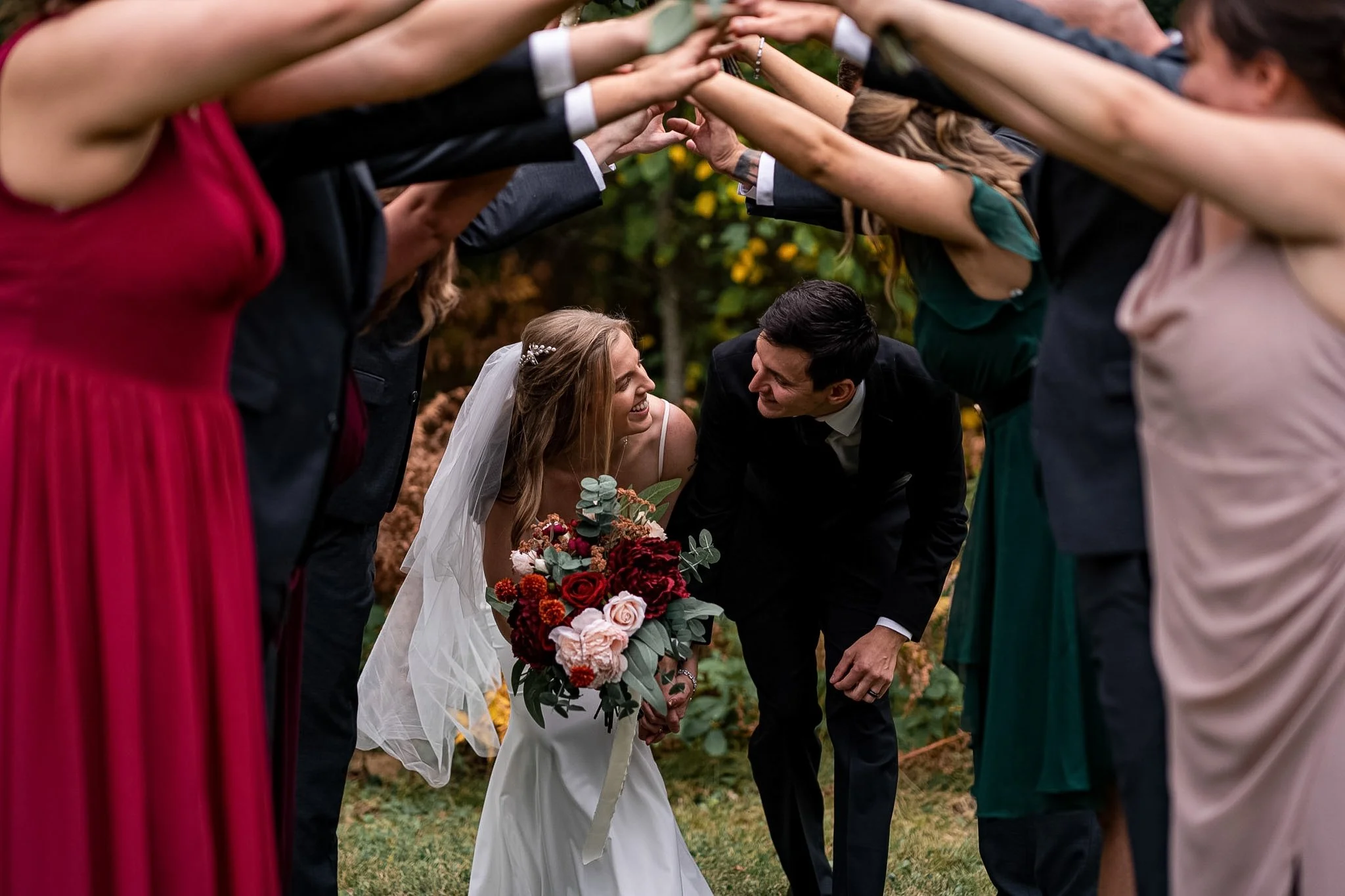 Bride and groom smiling and looking at each other, surrounded by friends and family forming a tunnel with their arms at a wedding outdoors.