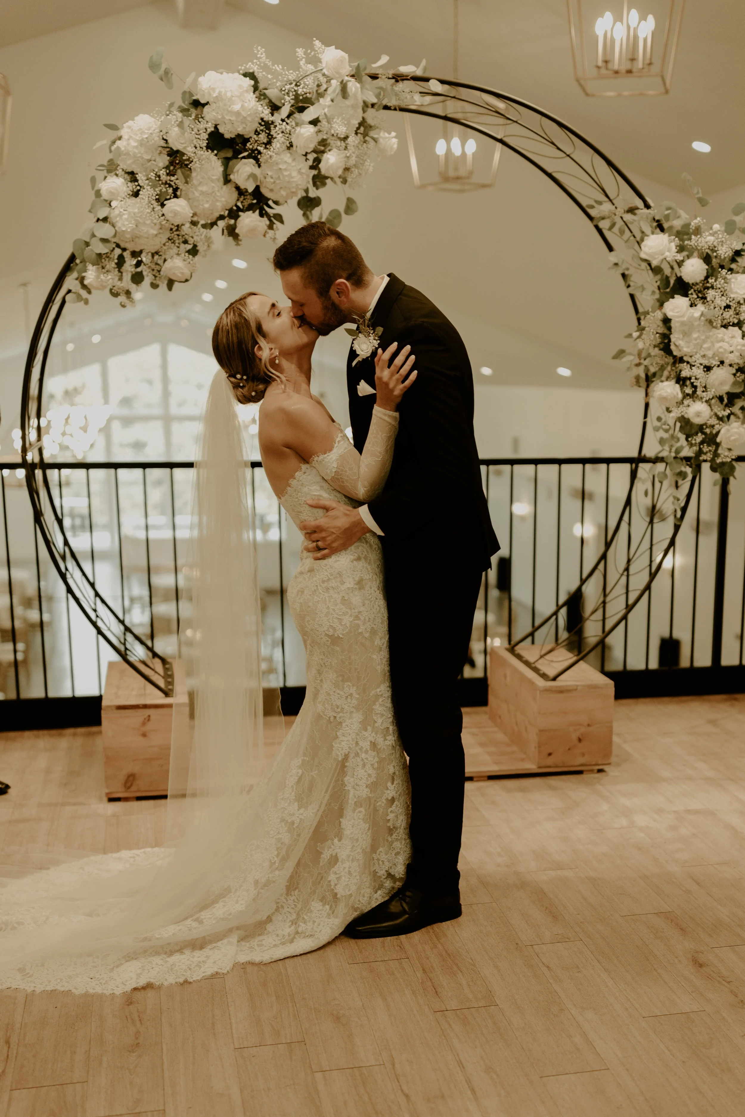 A bride and groom sharing a kiss at their wedding, standing under a circular floral arch decorated with white flowers, indoors with soft lighting.