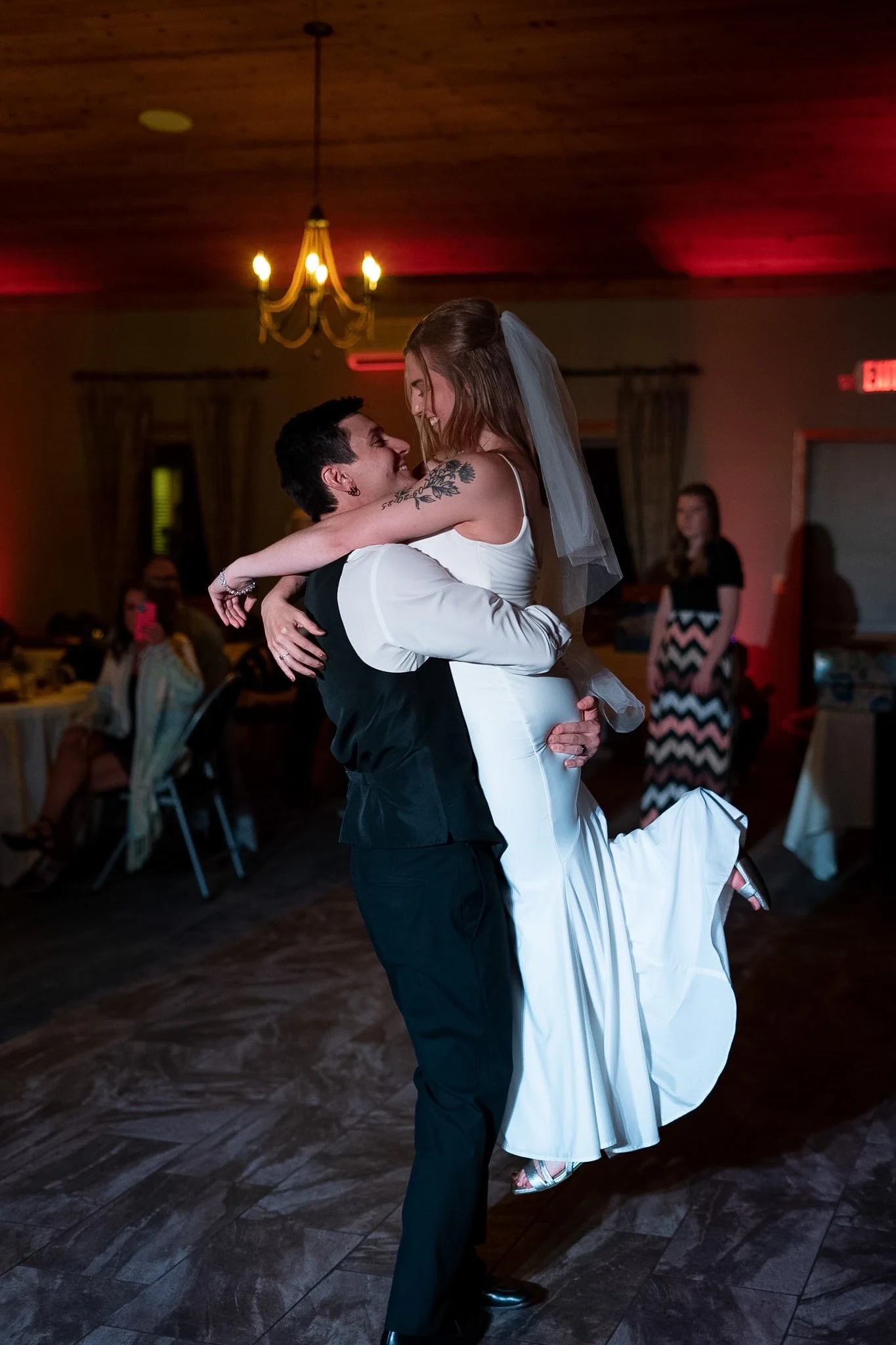A couple dancing at a wedding reception, with the groom lifting the bride off the ground. The bride is wearing a white wedding dress and veil, and the groom is in a black vest and trousers. Guests are seated at tables in the background, and a woman i