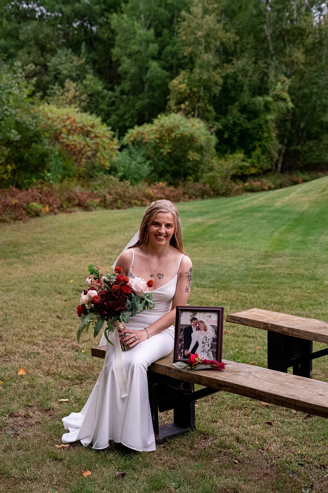 A smiling bride in a white wedding dress sits on a wooden bench outdoors, holding a bouquet of red, pink, and white flowers. Next to her on the bench is a framed wedding photo and a red flower.