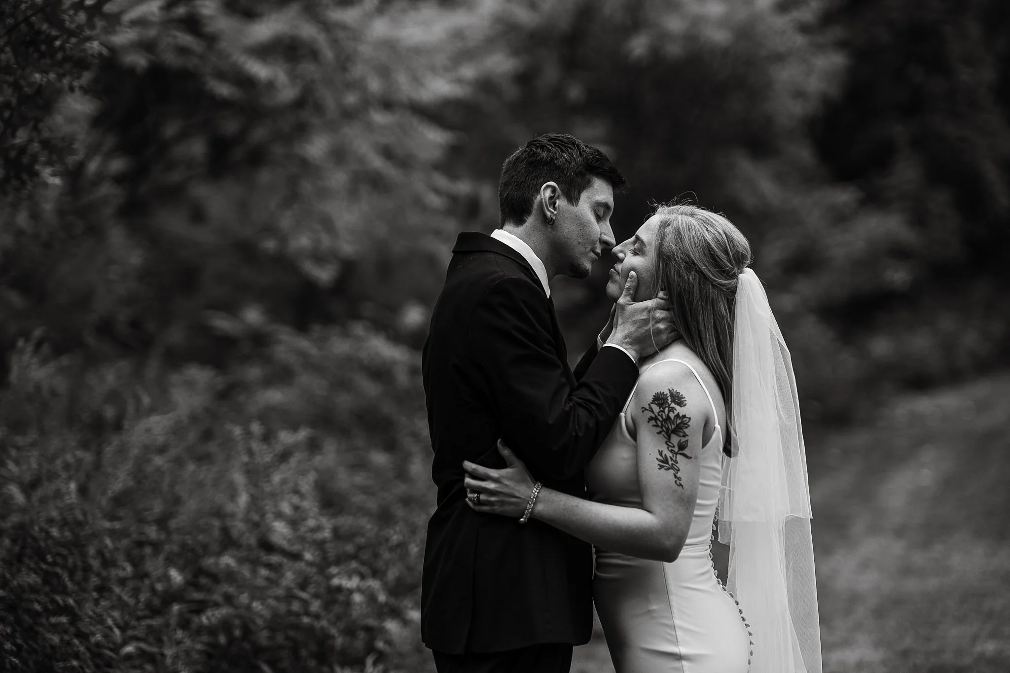 A couple on their wedding day, the groom gently holding the bride's face, both with closed eyes, outdoors with blurred trees in the background, in black and white.