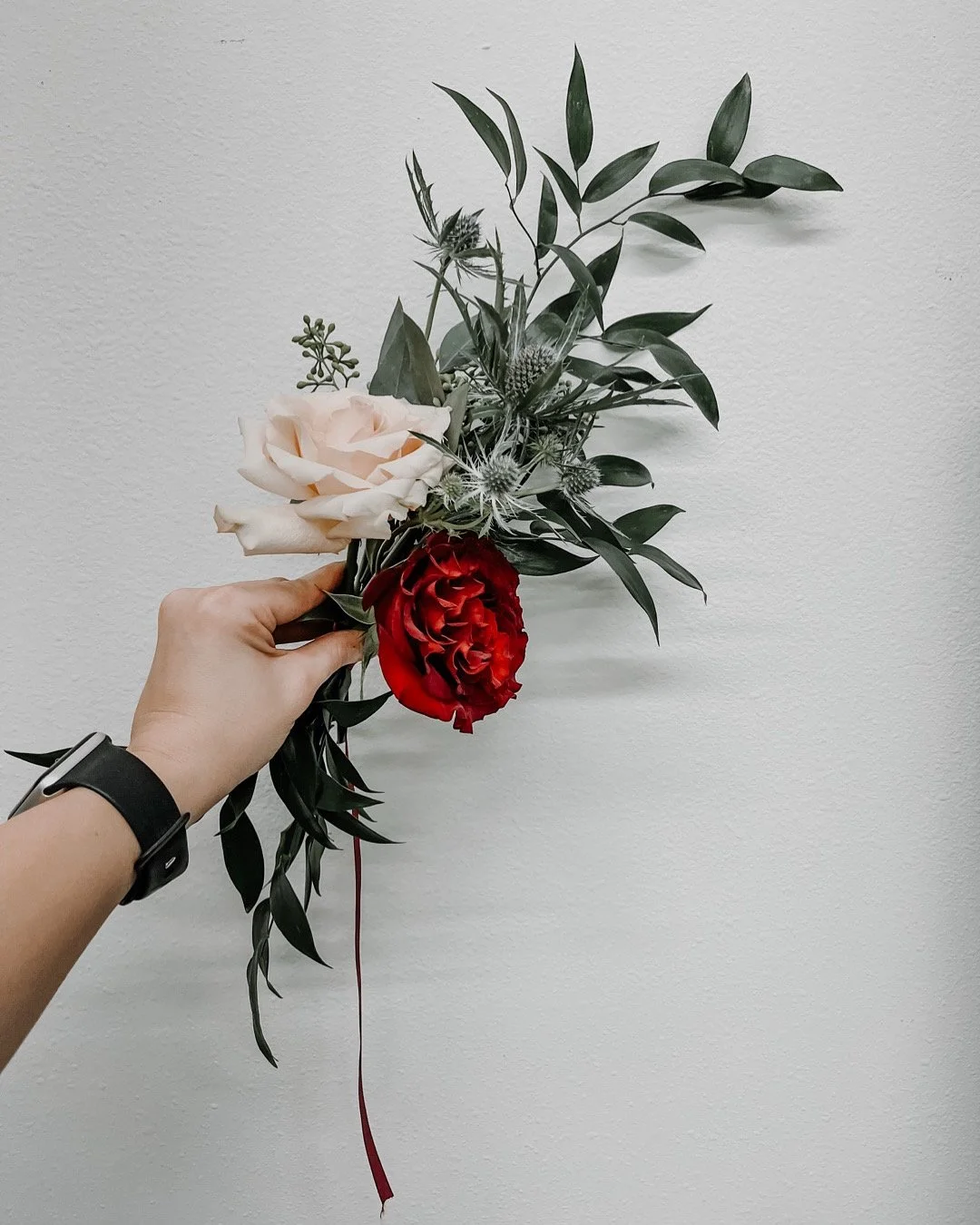 A hand holding a floral arrangement with a light pink rose, a red peony, and various green leaves, set against a plain white wall.
