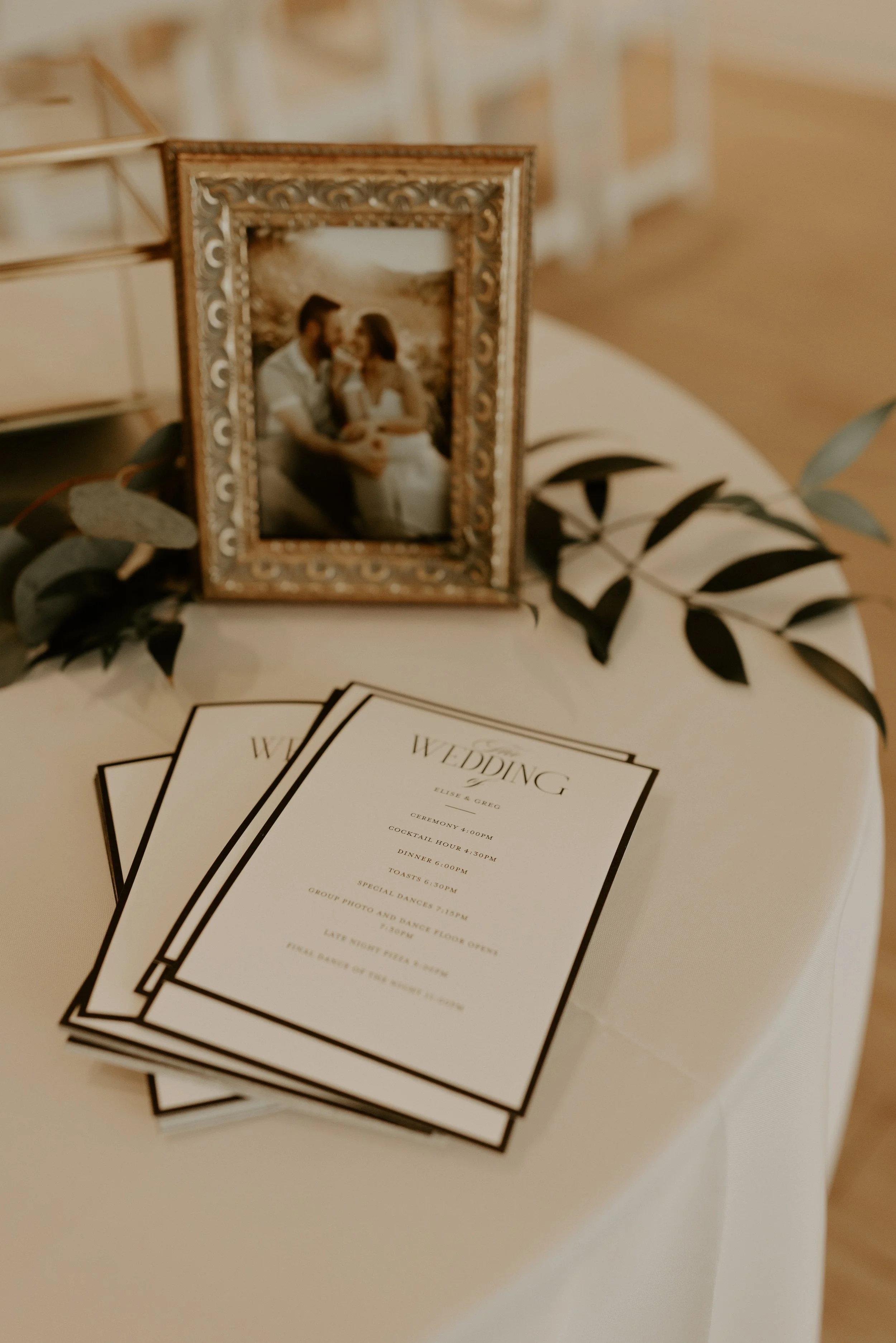 A framed photograph of a couple at a wedding, placed on a table alongside wedding programs, with greenery as decoration.