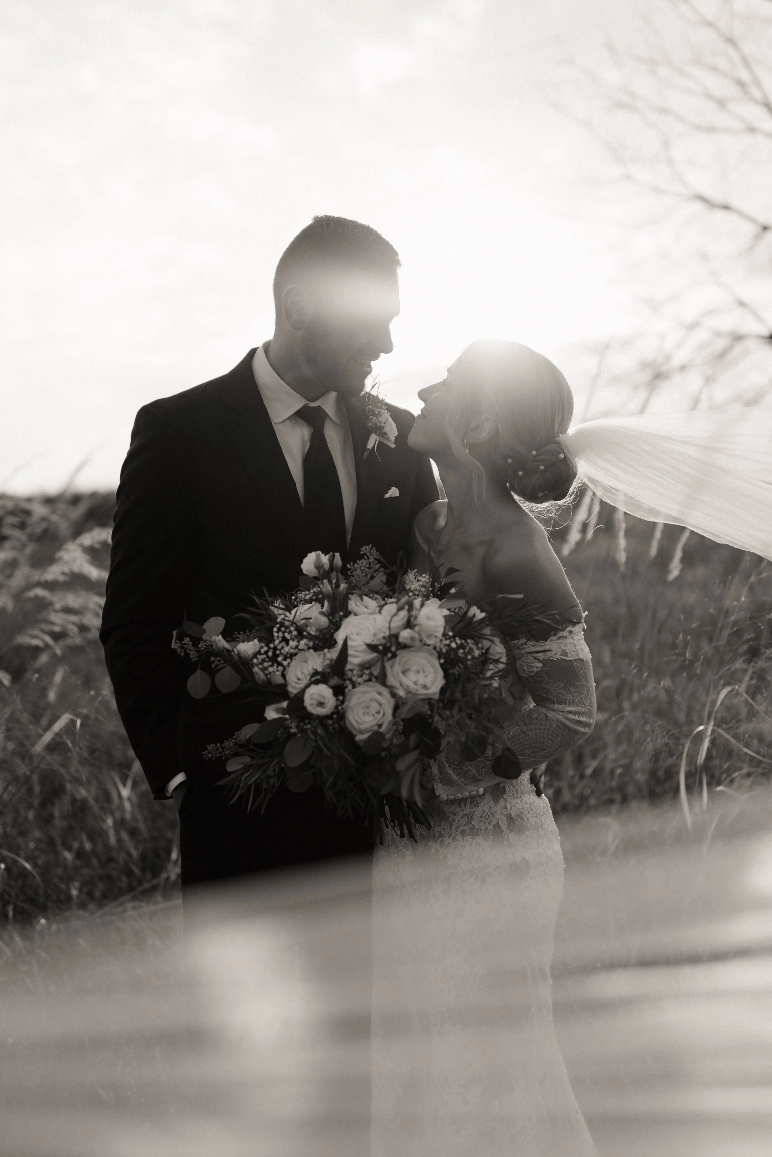 A black and white photo of a bride and groom standing close together outdoors at sunset, with the bride holding a bouquet of flowers, and the couple gazing affectionately at each other.