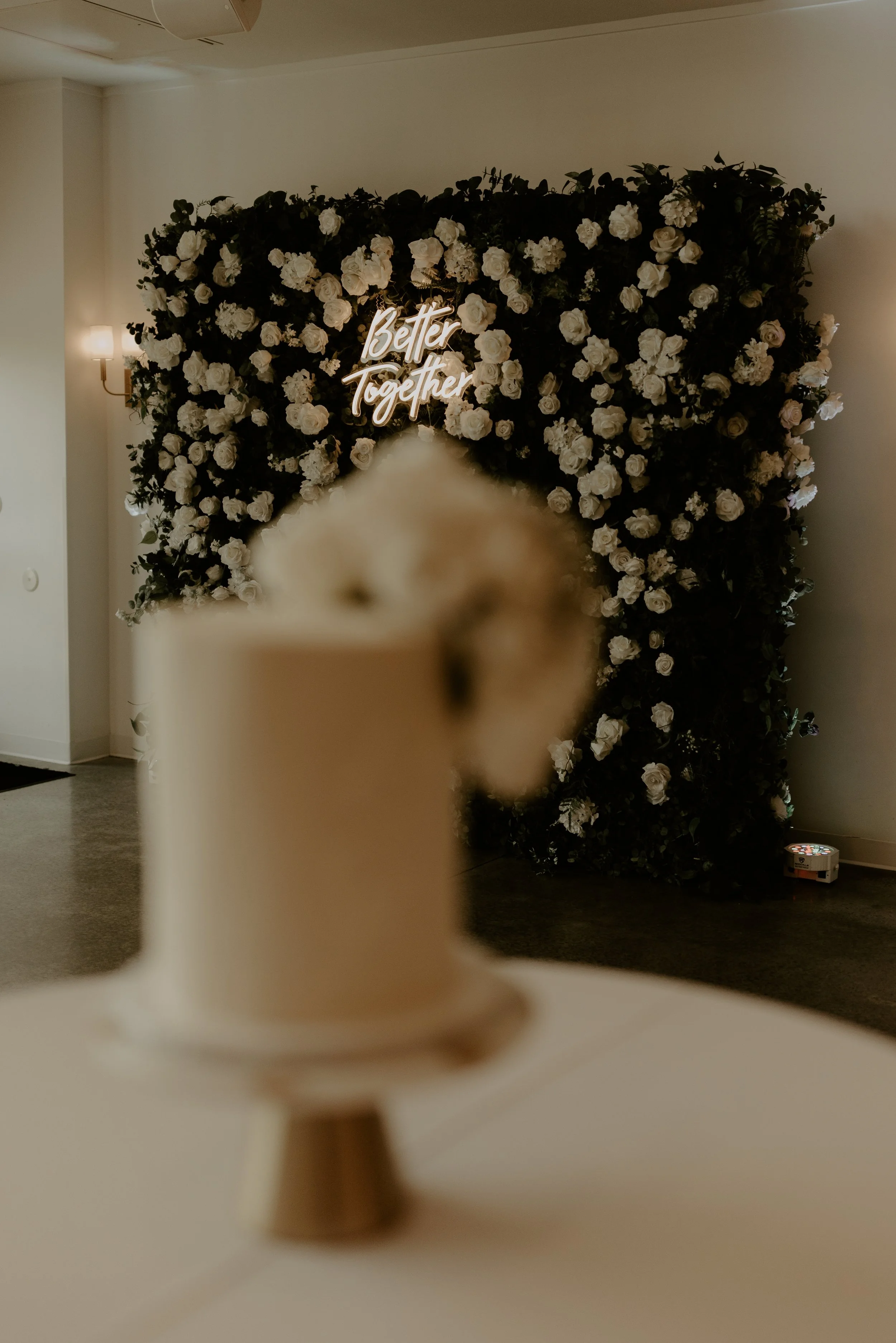 Blurred beige cup of coffee or tea with a black straw on a white table, in front of a floral backdrop with white roses and a neon sign that says 'Better Together'