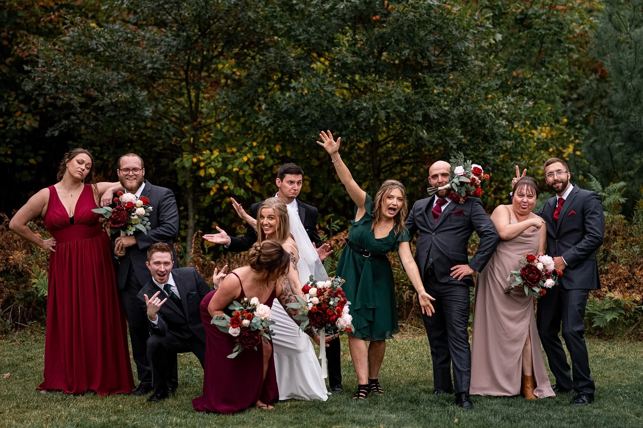 A group of nine people, dressed in formal wedding attire, are posing outdoors with a backdrop of trees with autumn leaves. Some are smiling and making playful gestures.