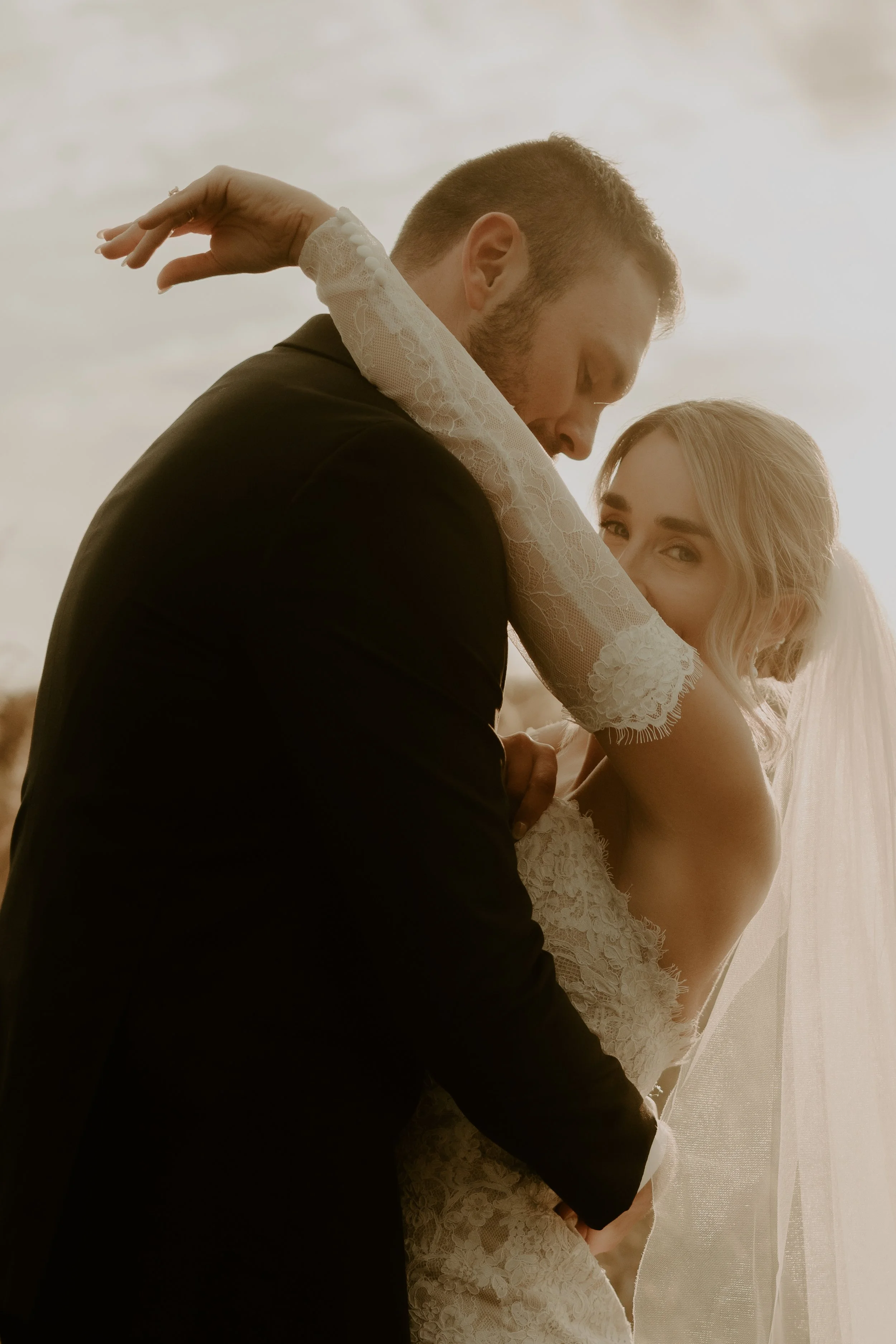 A bride and groom embracing on their wedding day, with the bride wearing a lace wedding dress and veil, and the groom in a black suit, against a soft, natural backdrop.
