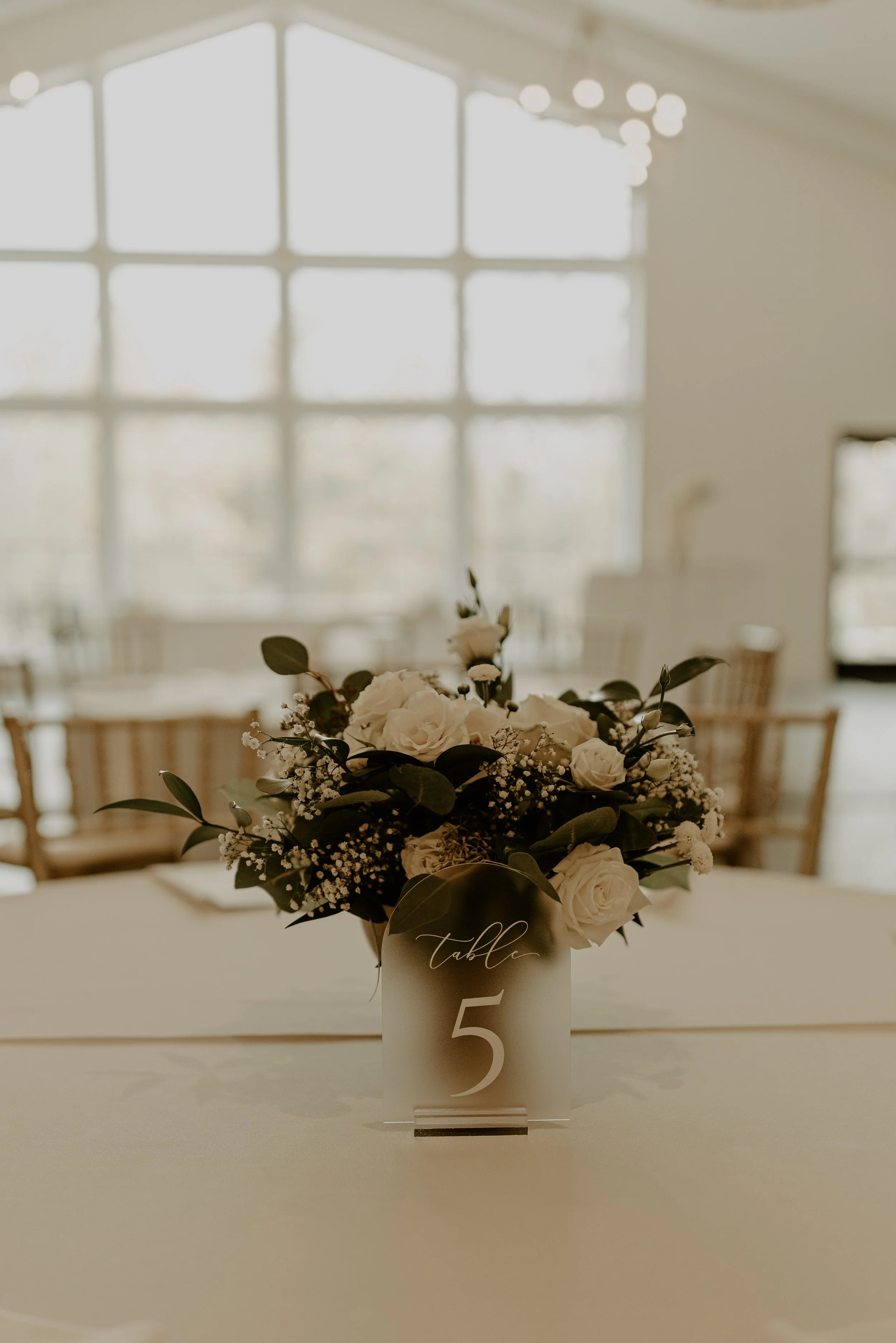 Floral centerpiece with white roses and greenery on a table at a wedding reception, with a table number '5' sign.