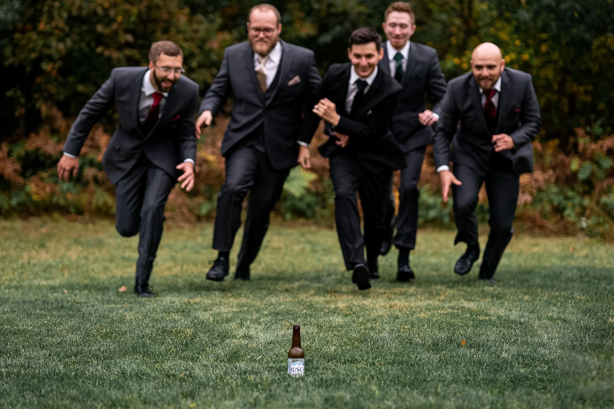 A group of five men in tuxedos running towards a beer bottle on a grassy field.