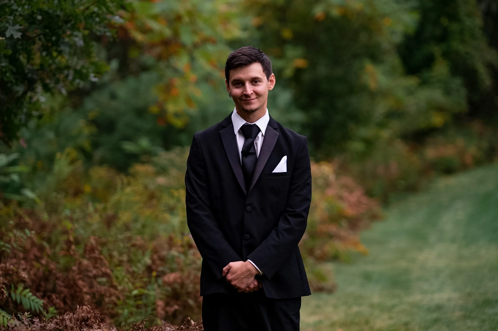 A young man dressed in a black tuxedo with a white shirt and black tie, standing outdoors in a forested area with green foliage and autumn-colored leaves.