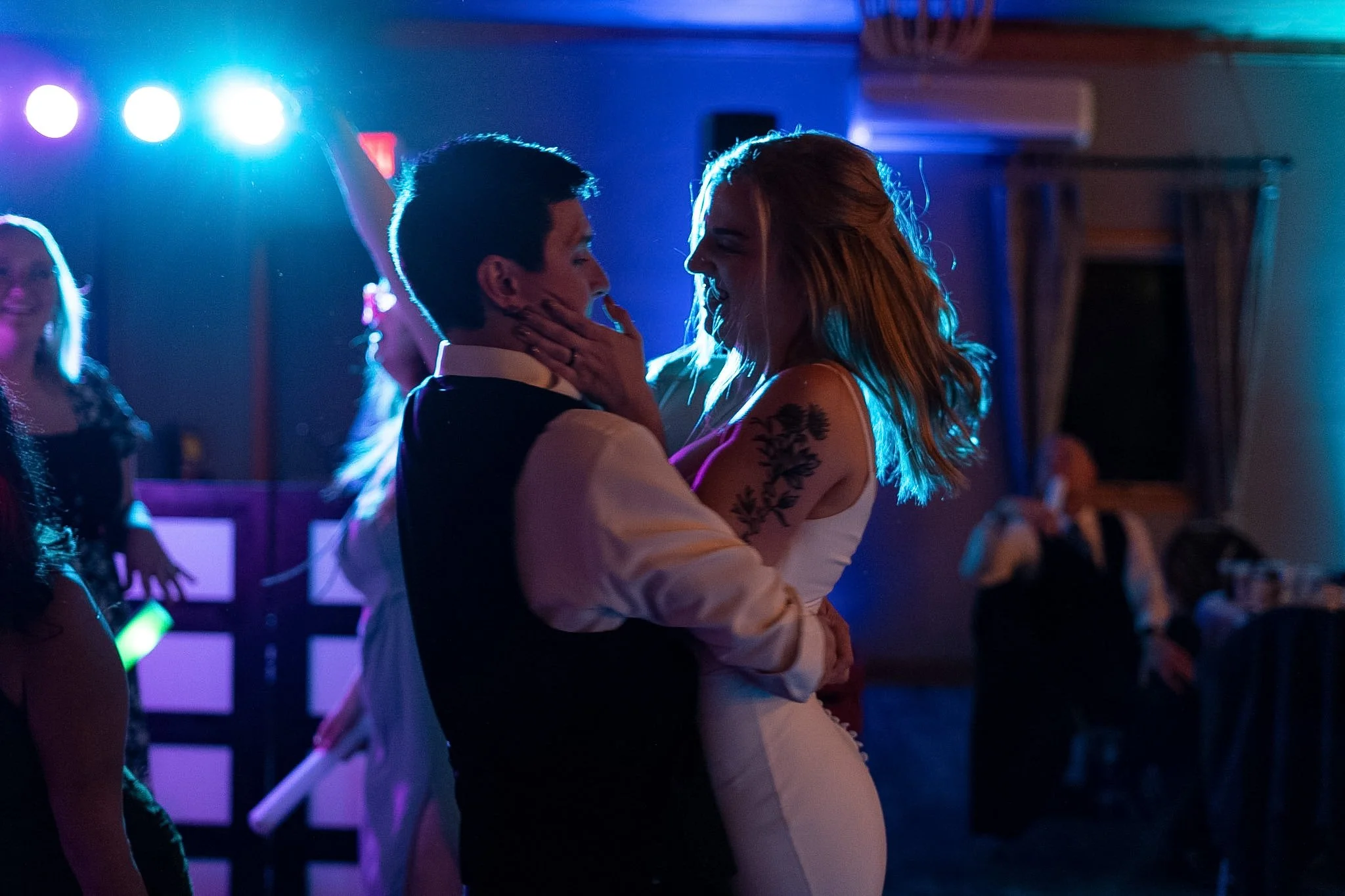 A couple dancing closely at a party or wedding reception, with colorful party lights in the background.
