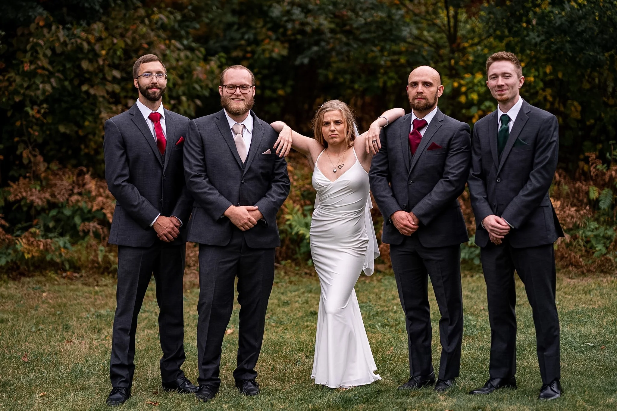 A woman in a white dress stands with four men in gray suits and ties, outdoors on grass with trees in the background. The woman has her arms over two men's shoulders and looks slightly displeased, while the men stand with their hands clasped in front