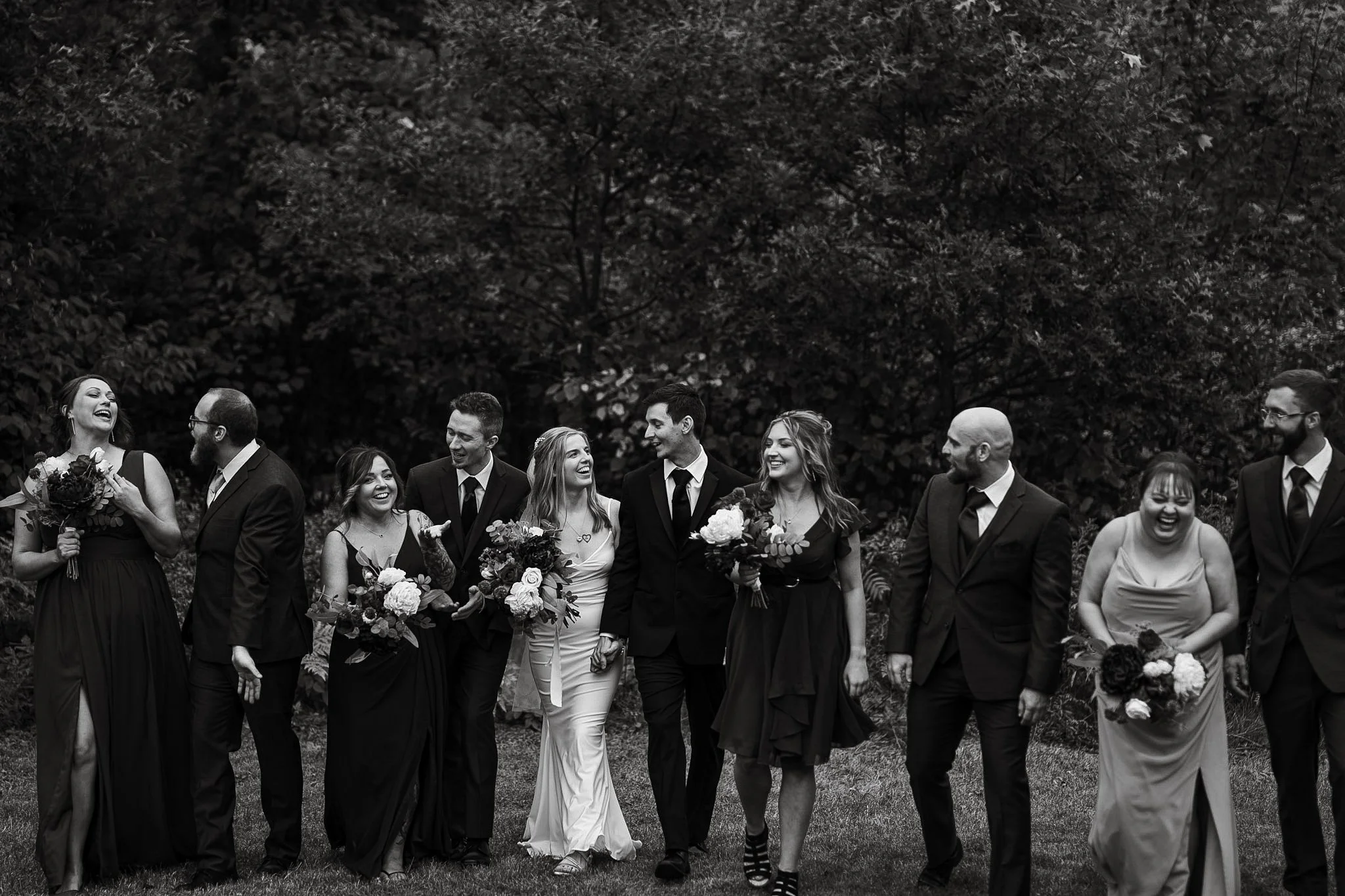 Black and white photo of a diverse group of people, dressed in formal attire, standing outdoors on grass with trees in the background, smiling and laughing together at a celebration or wedding.