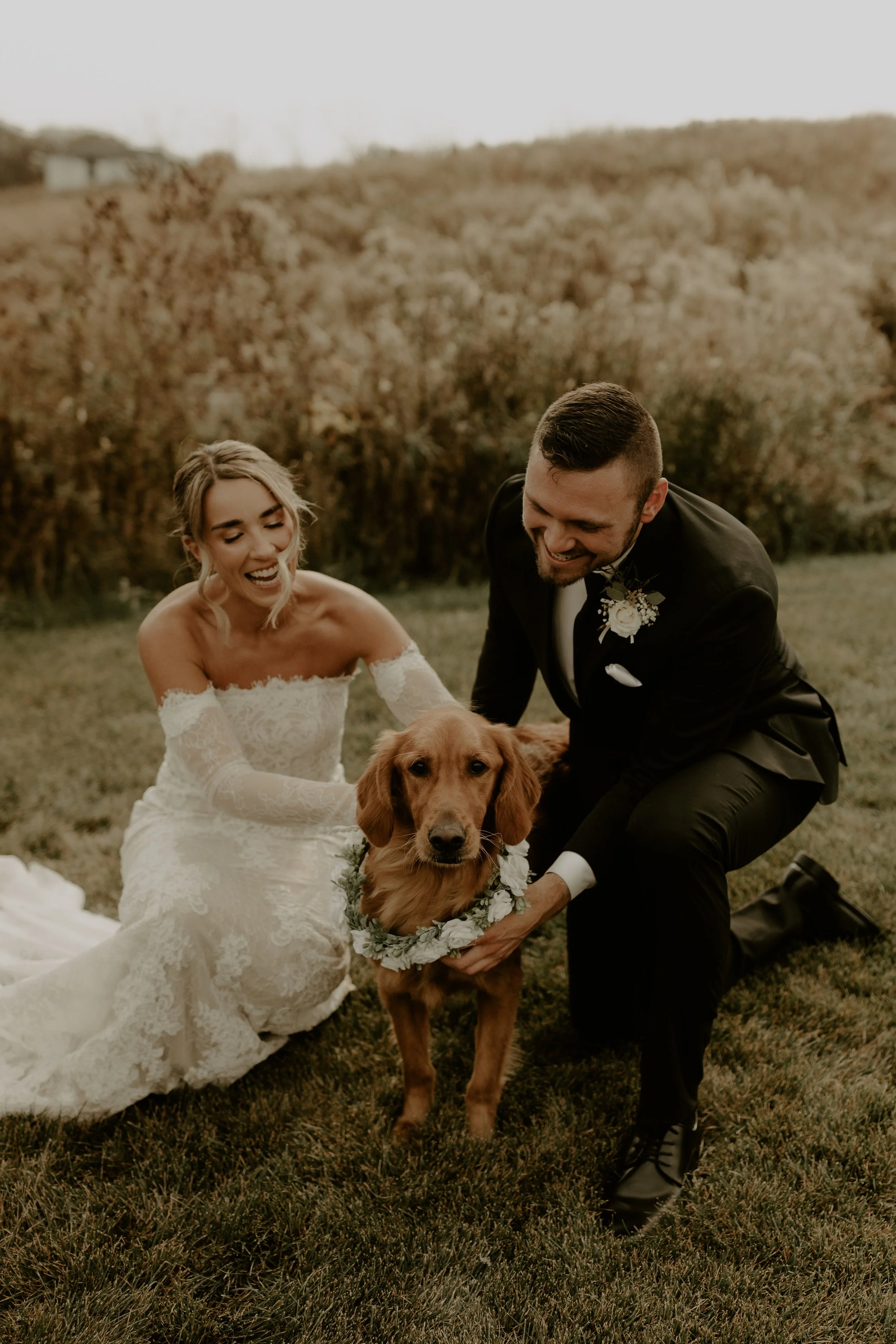 A bride and groom kneeling on grass, holding a golden retriever dog with a floral collar, outdoors during their wedding.