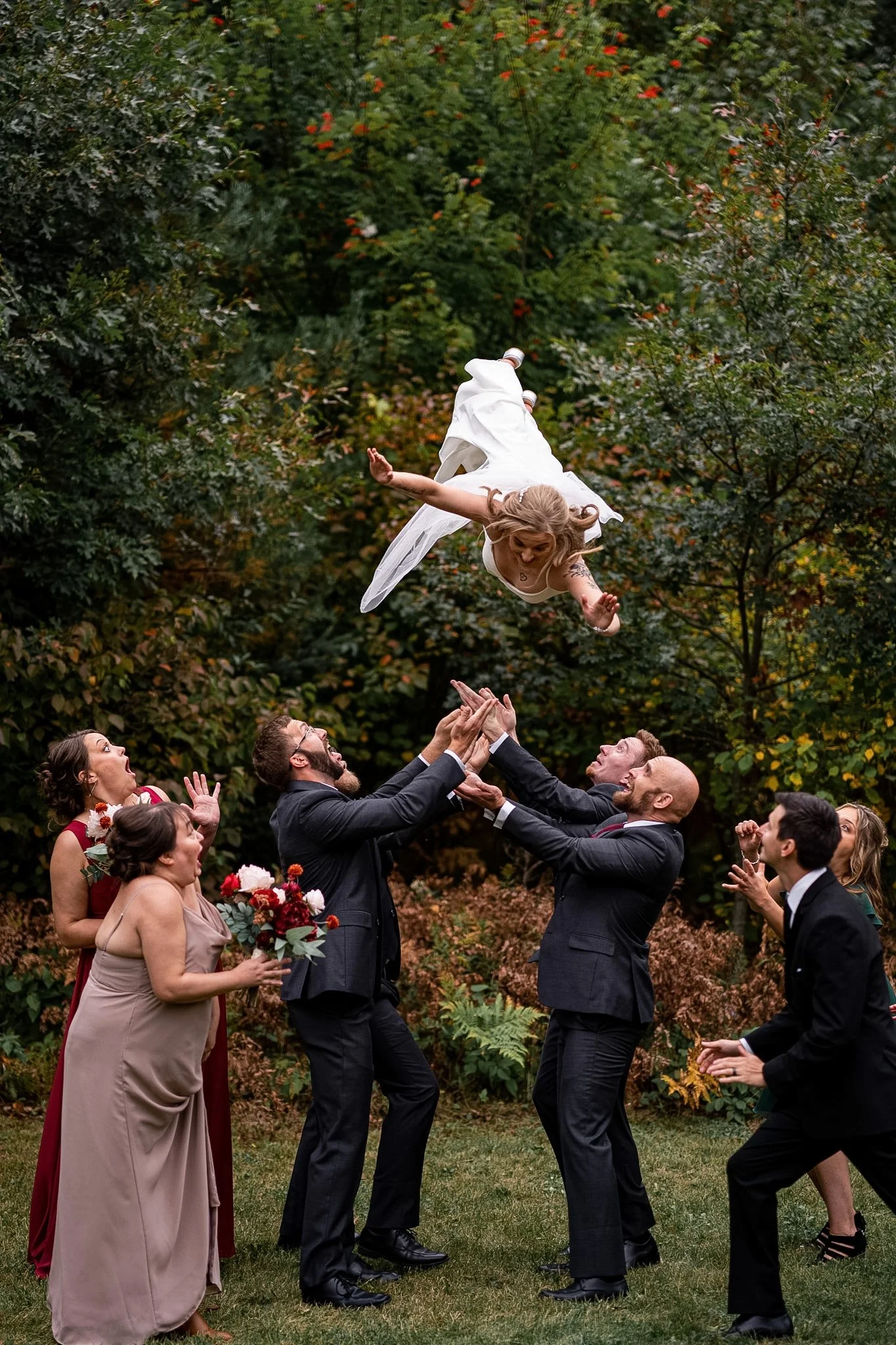 A bride in a white dress is being tossed into the air by a group of people at an outdoor wedding reception, with trees and foliage in the background.