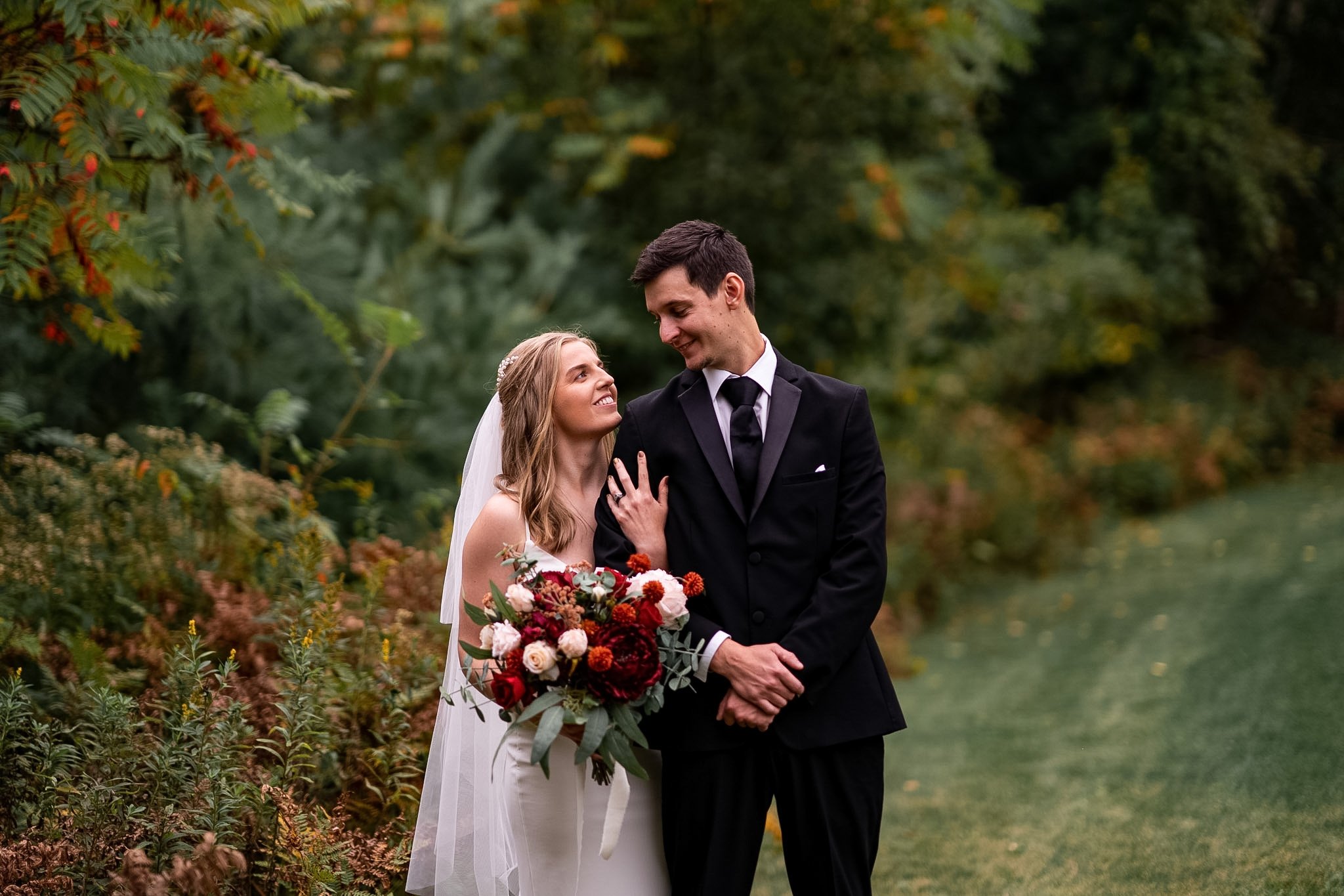 A bride and groom sharing a moment outdoors with greenery and trees in the background. The bride is holding a bouquet of red, white, and pink flowers, and the groom is dressed in a black suit. They are smiling and looking at each other.