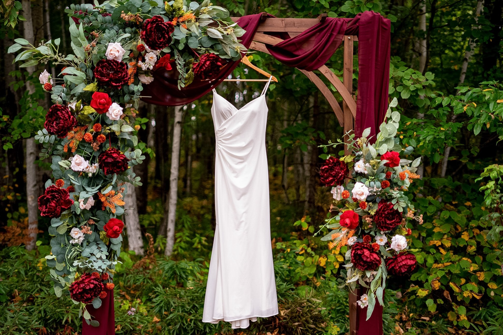 Wedding dress hanging on a wooden arch decorated with burgundy and light pink flowers and draped burgundy fabric, set in a forested outdoor area.