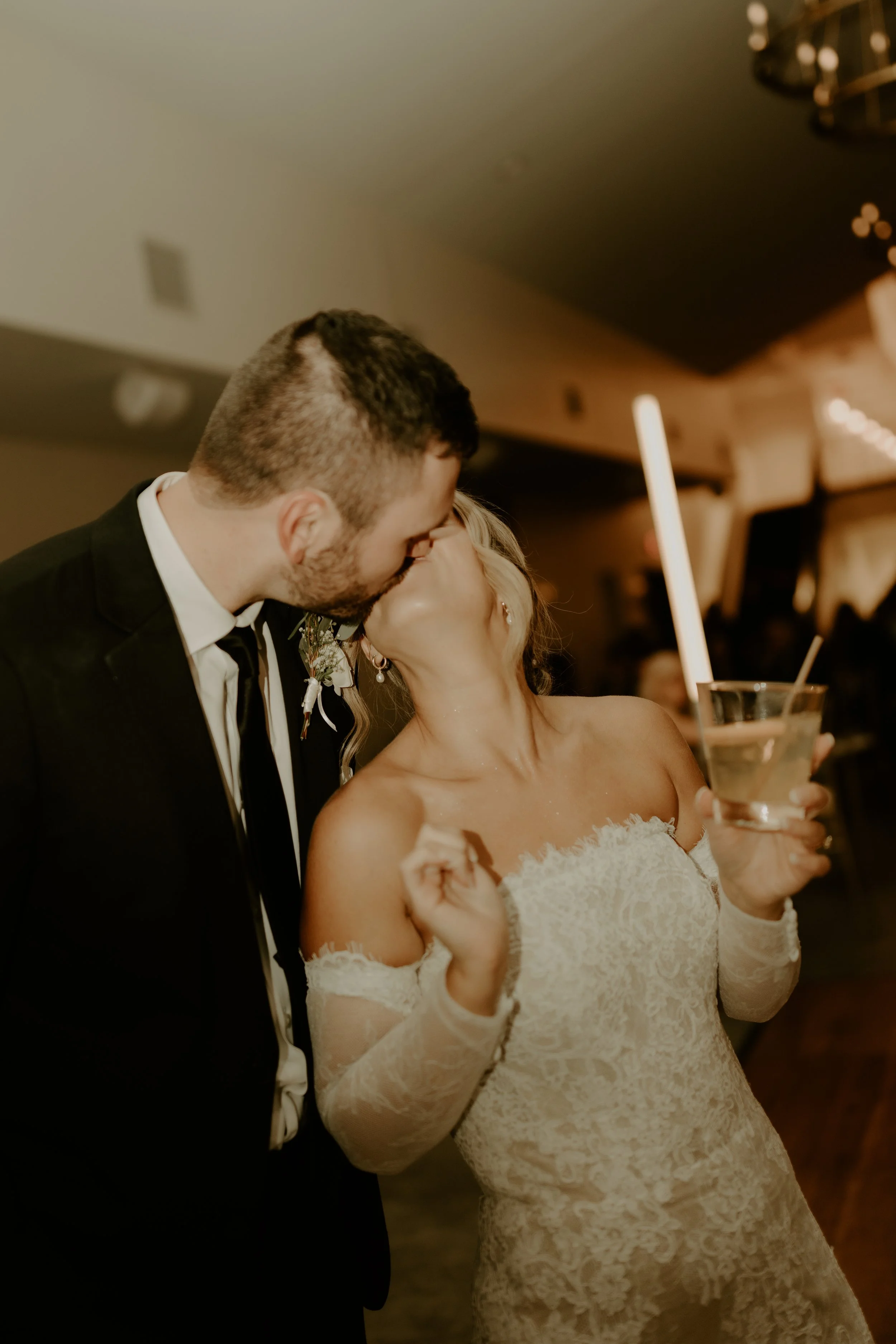 A newlywed couple sharing a kiss at their wedding reception, with the bride holding a drink and wearing an off-shoulder lace wedding dress.