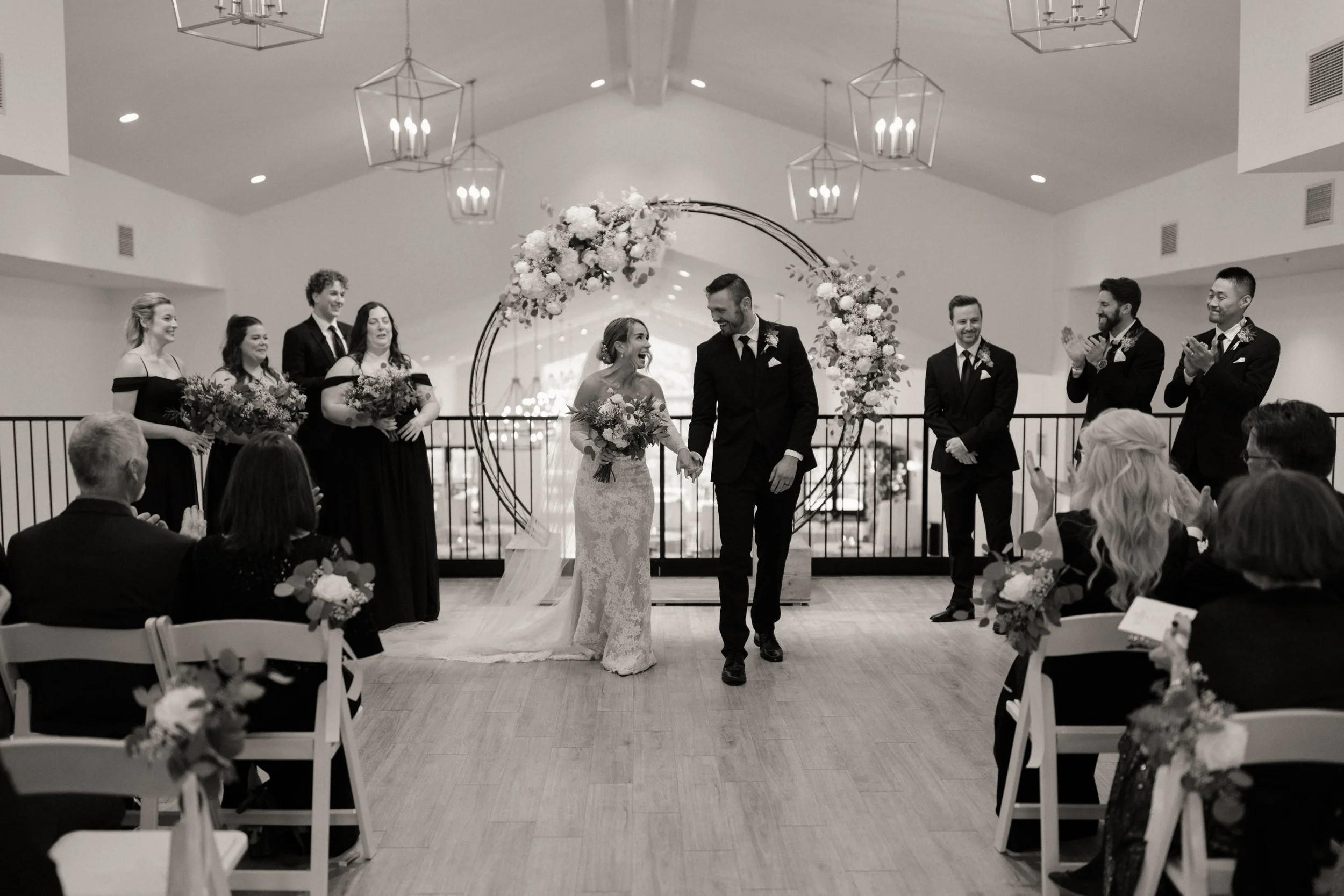 A black and white photo of a wedding ceremony. The bride and groom are holding hands and smiling at each other, walking forward. The bride is in a lace wedding dress, and the groom is in a suit. Behind them, bridesmaids and groomsmen are clapping and