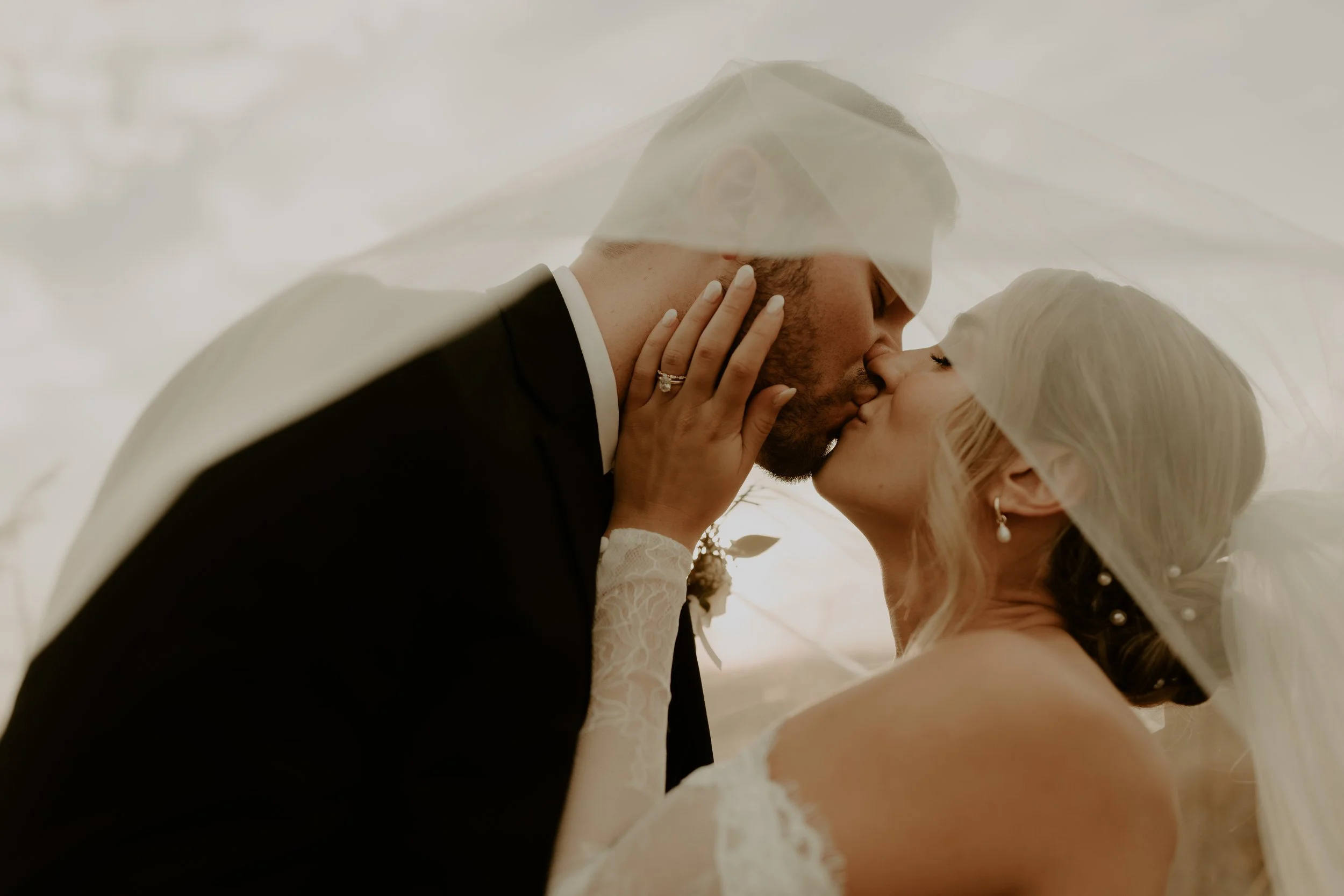 A bride and groom share a kiss beneath a wedding veil, with the bride's hand gently touching the groom's face.